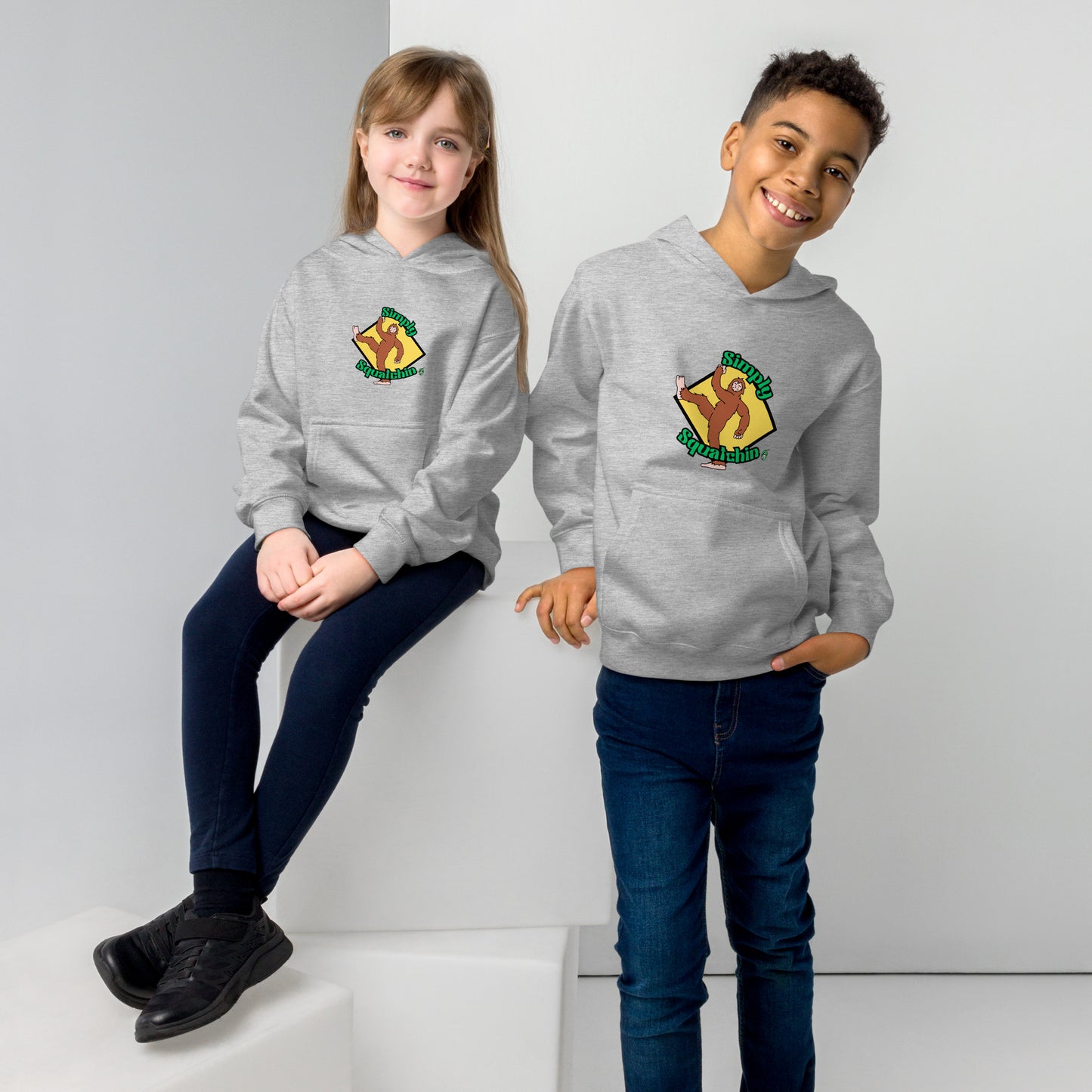 A boy standing and a girl sitting both wearing a grey kids graphic hoodie with a brown sasquatch and the statement, Simply Squatchin on a yellow background, by Wizard Prints