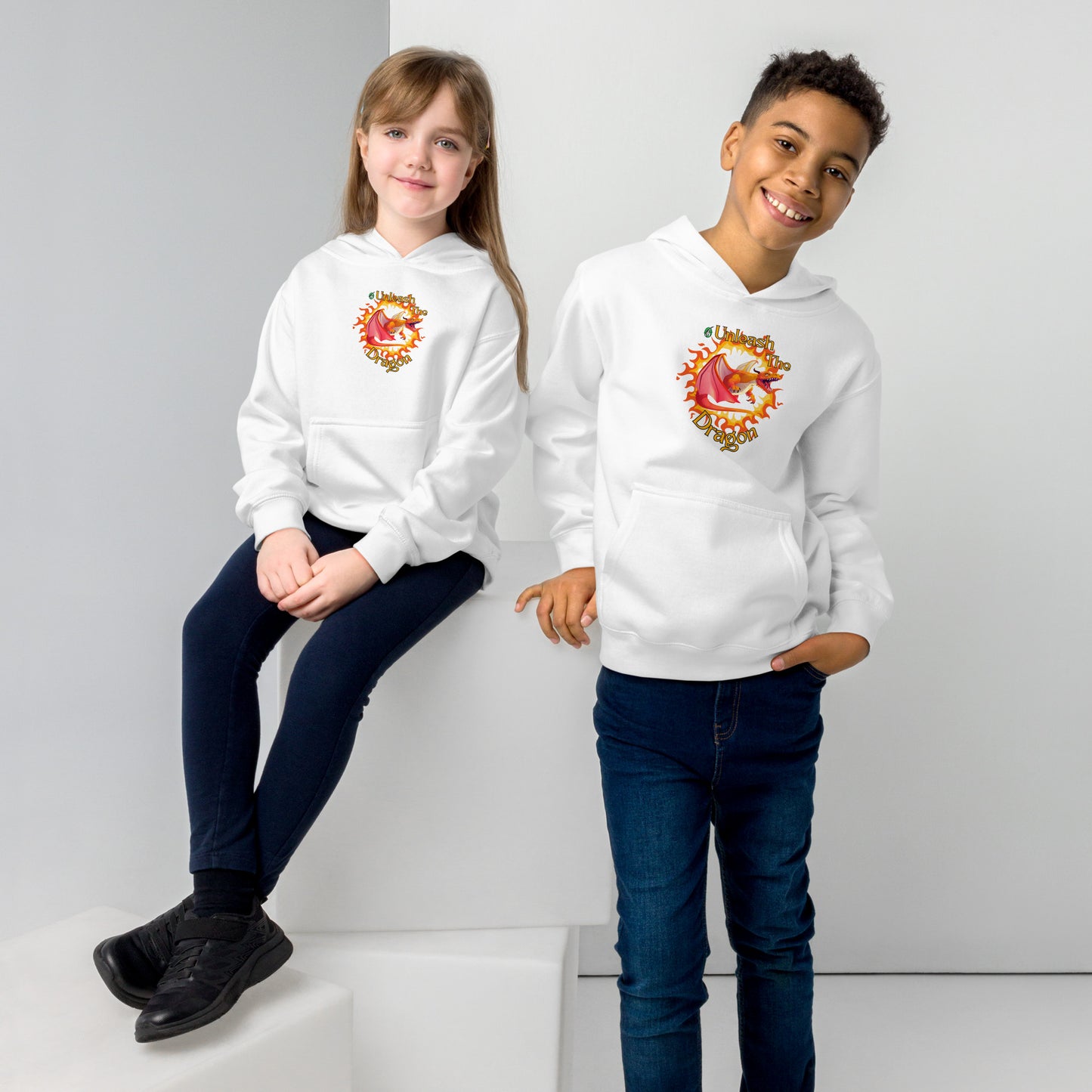 A boy standing and a girl sitting both wearing a white kids graphic hoodie with a red dragon breathing fire and the statement unleash the dragon, by Wizard Prints
