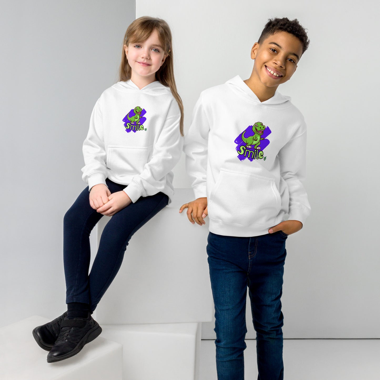 A boy standing and a girl sitting both wearing a white kids graphic hoodie with a green dinosaur and the word Smile on a purple background, by Wizard Prints