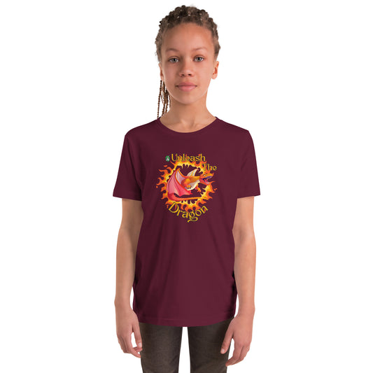 A girl standing wearing a maroon kids t-shirt with the printed graphic of a fire-breathing dragon with the statement, Unleash the Dragon. Designed by Wizard Prints