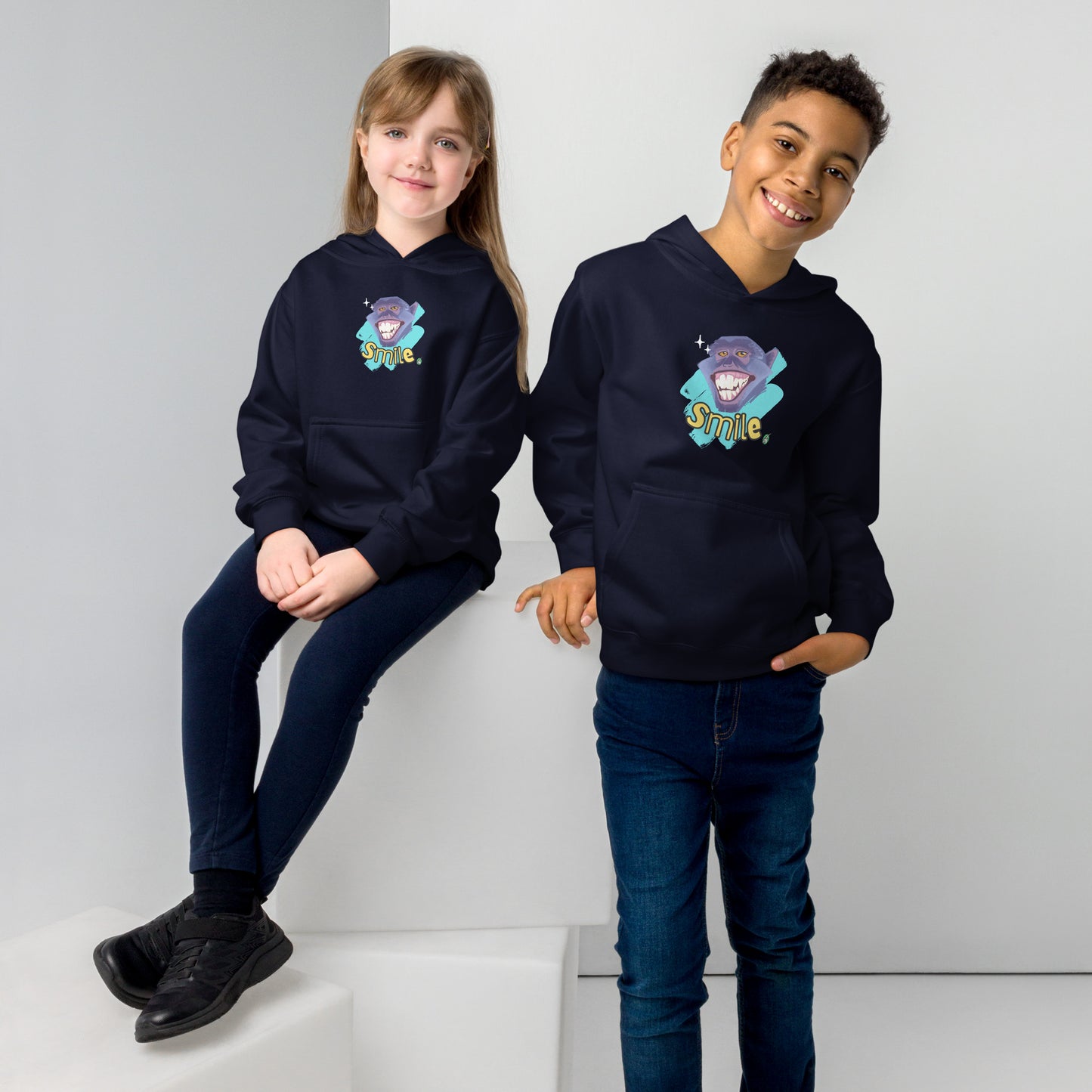 A boy standing and a girl sitting both wearing a navy blue kids graphic hoodie with a smiling monkey face and the word, Smile, by Wizard Prints