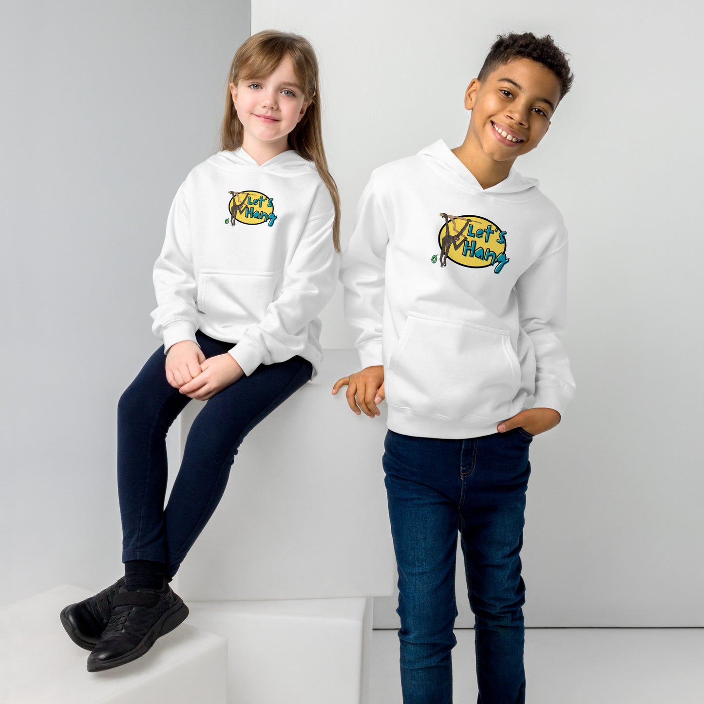 A boy standing and a girl sitting wearing a white hoodie with a funny graphic on the front of a monkey hanging from the tree branch saying, Let's Hang by Wizard Prints and jeans on a grey background