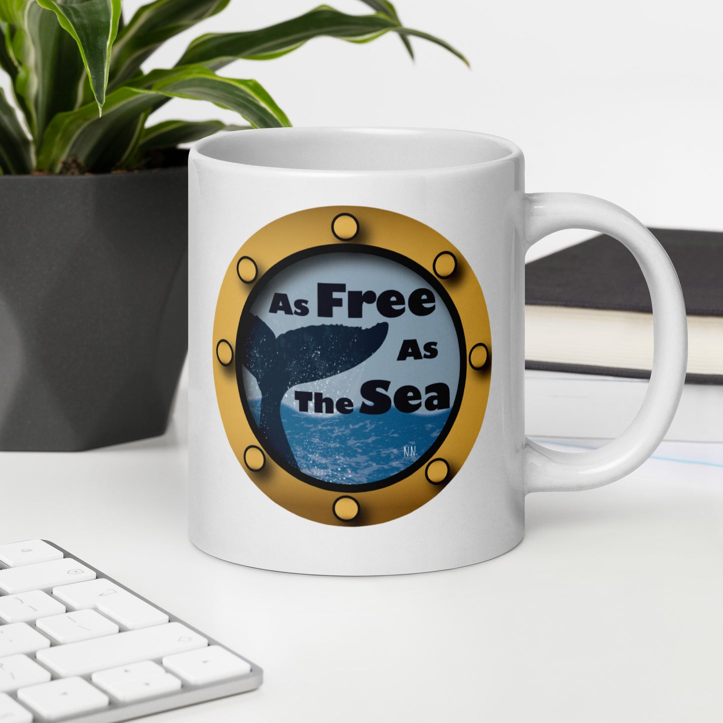 White mug with a whale and sea graphic and 'Free as the Sea' text on a desk with a plant and books in the background.