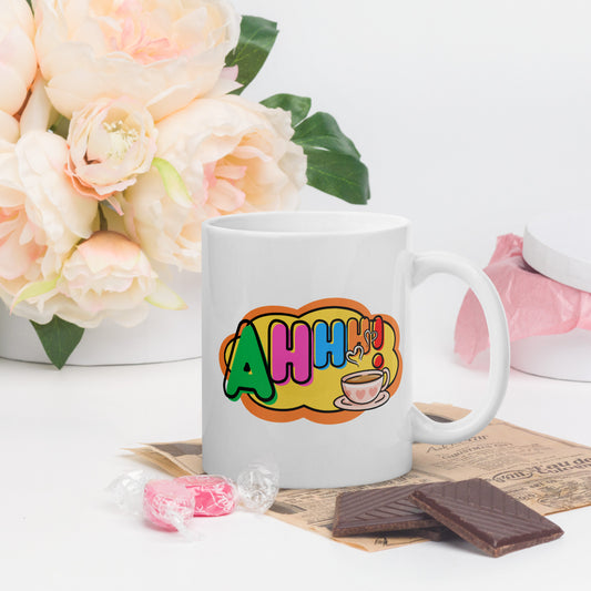 White mug with colourful design featuring a coffee cup and 'Ahhh!' text, placed on a surface with flowers and snacks.