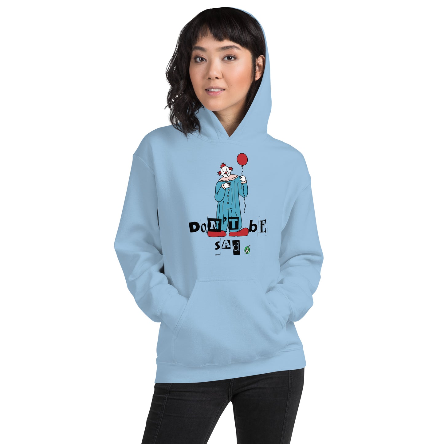 A woman standing and wearing a baby blue hoodie with a printed graphic of a clown holding a balloon with the words, 'Don't Be Sad'. Designed by Wizard Prints.