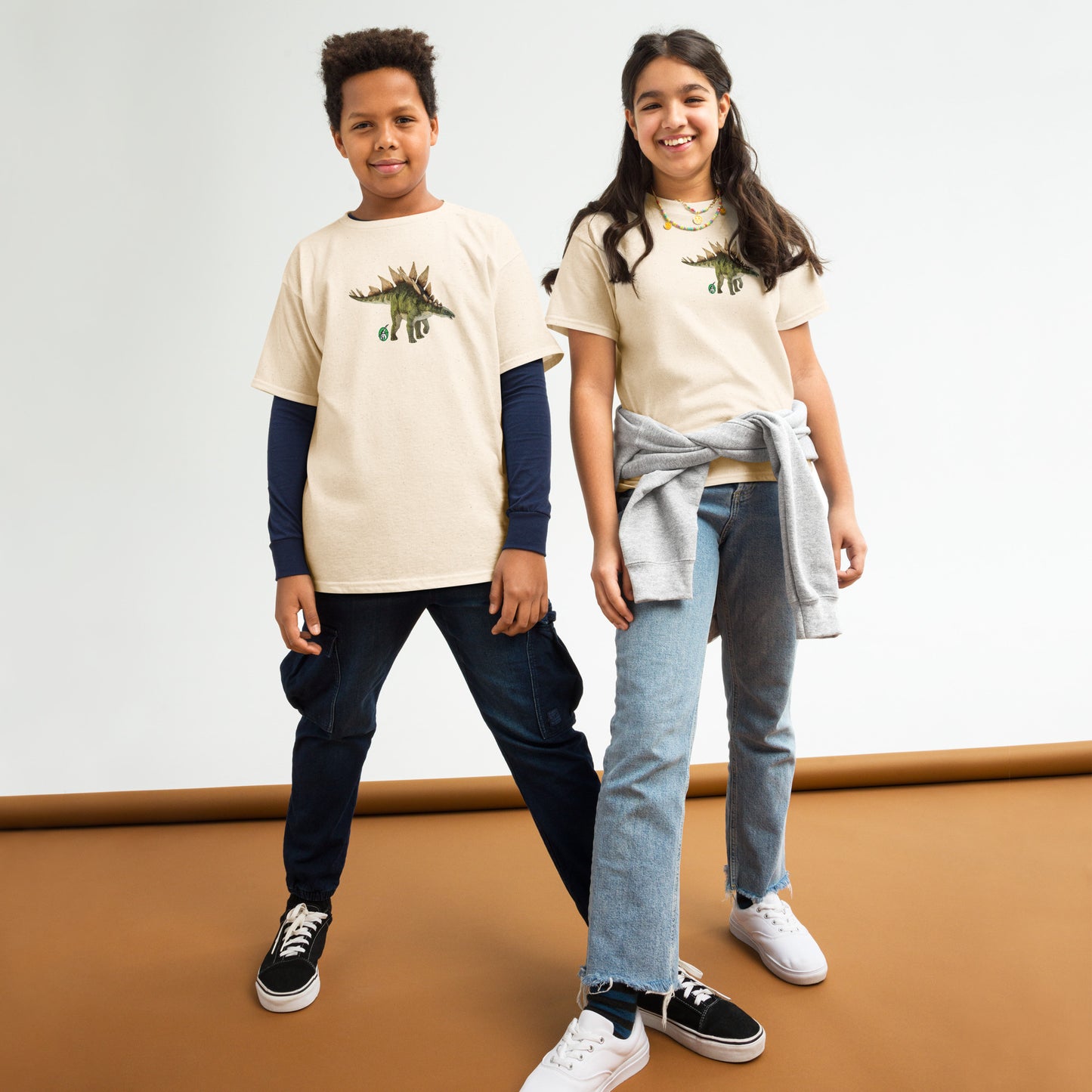 A boy and a girl standing and wearing beige t-shirts featuring a printed graphic of a life like stegosaurus, designed by Wizard Prints.