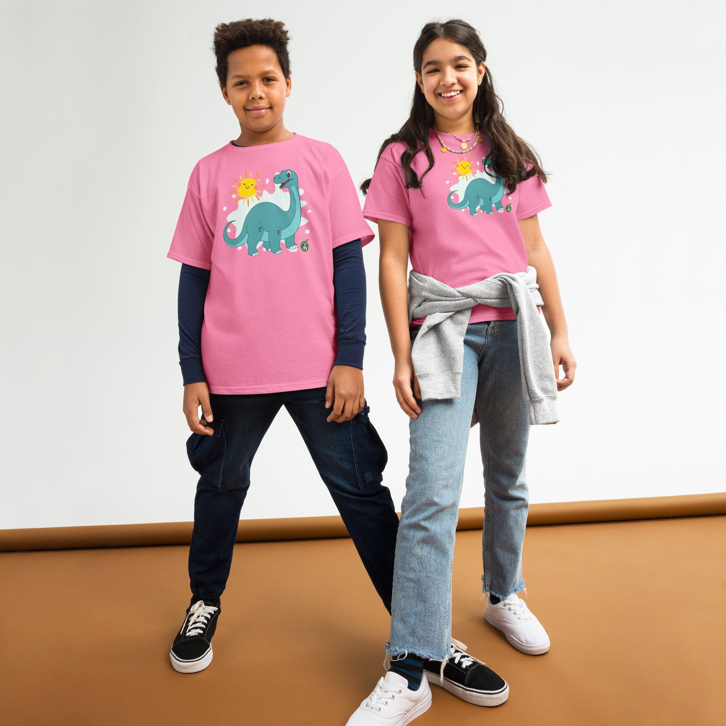 A boy and a girl standing and wearing pink t-shirts with a printed graphic of a smiling cartoon brachiosaurus and the sun. Designed by Wizard Prints.