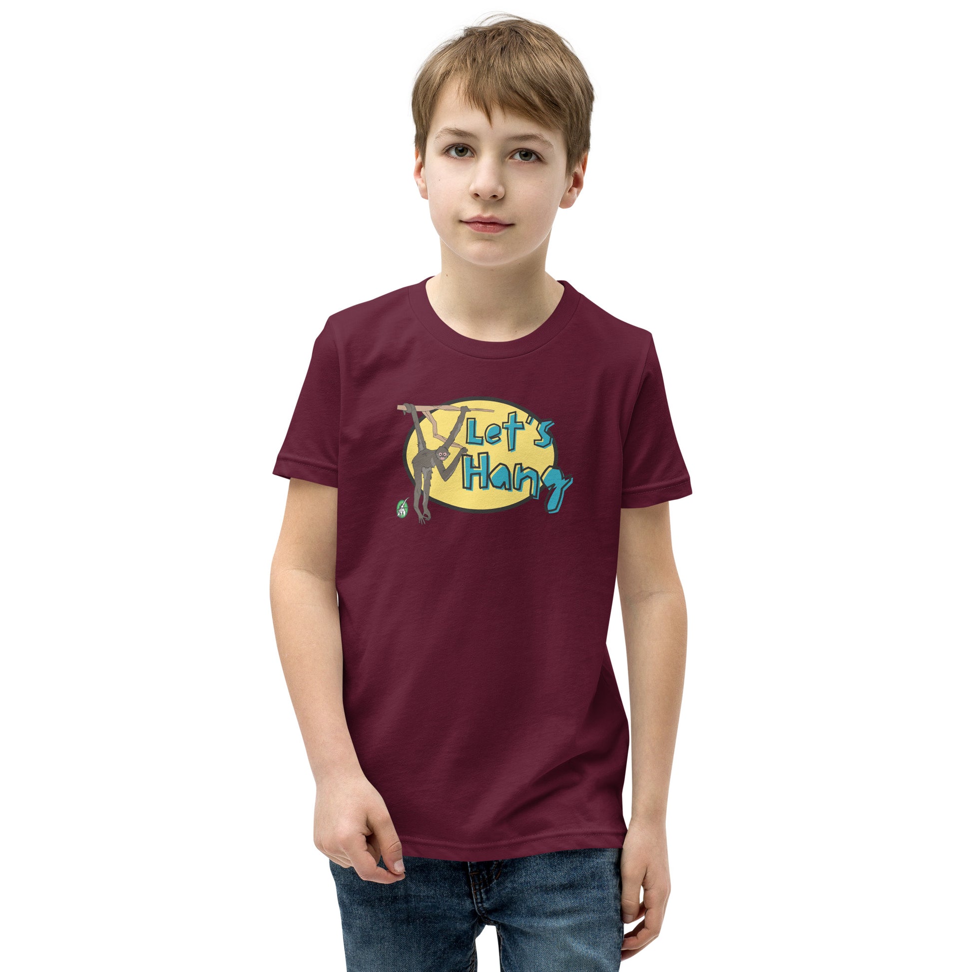 A boy standing wearing a dark red graphic tee shirt with a funny graphic on the front of a monkey hanging from the tree branch saying, Let's Hang by Wizard Prints and jeans on a white background
