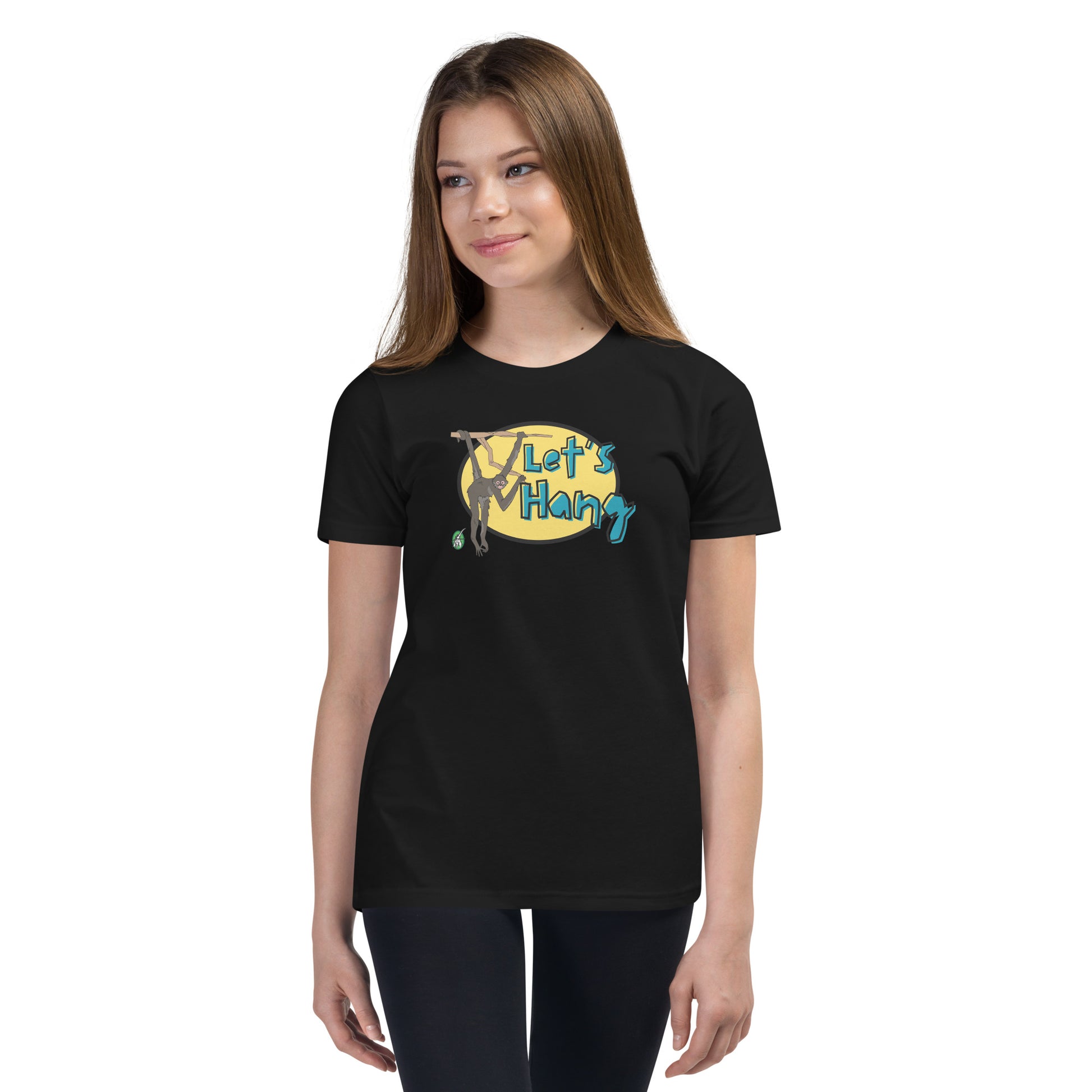 A girl standing wearing a black graphic tee shirt with a funny graphic on the front of a monkey hanging from the tree branch saying, Let's Hang by Wizard Prints and jeans on a white background
