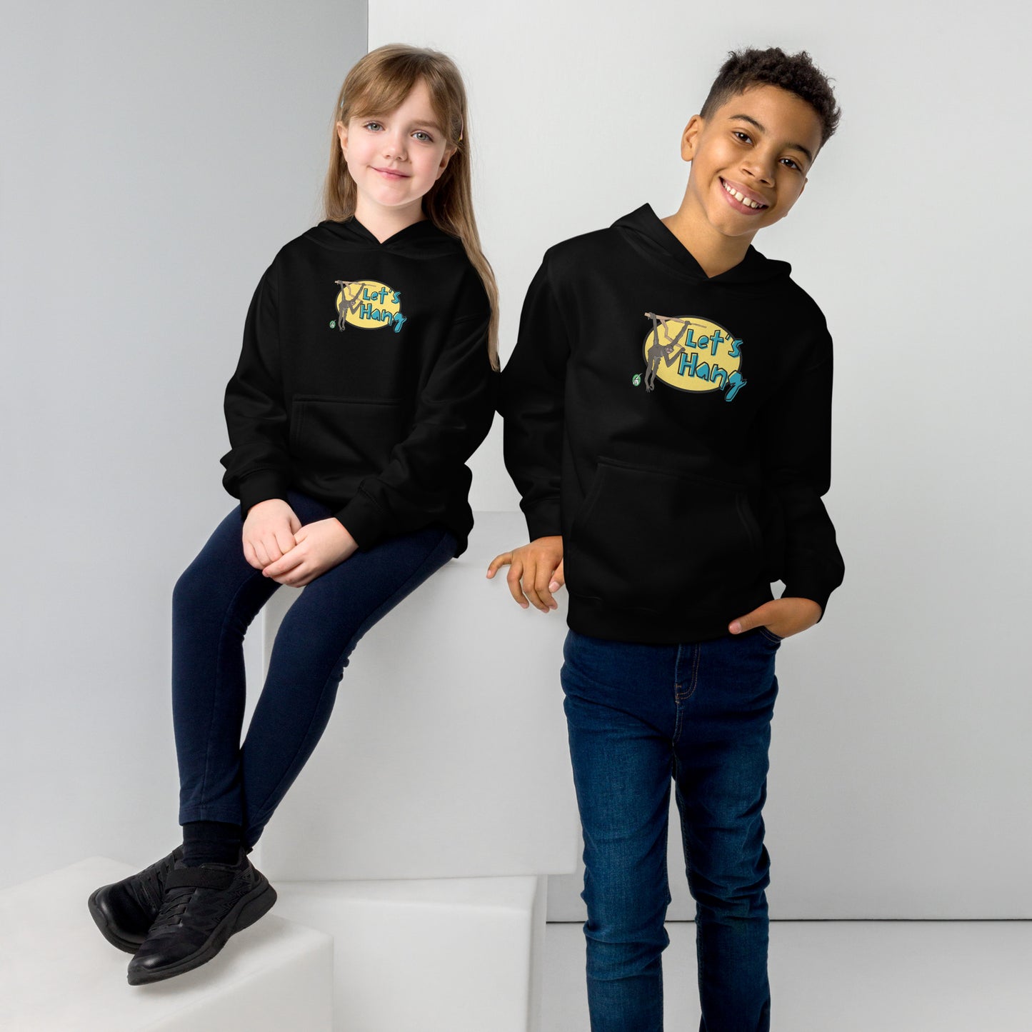 A boy standing and a girl sitting wearing a black hoodie with a funny graphic on the front of a monkey hanging from the tree branch saying, Let's Hang by Wizard Prints and jeans on a grey background