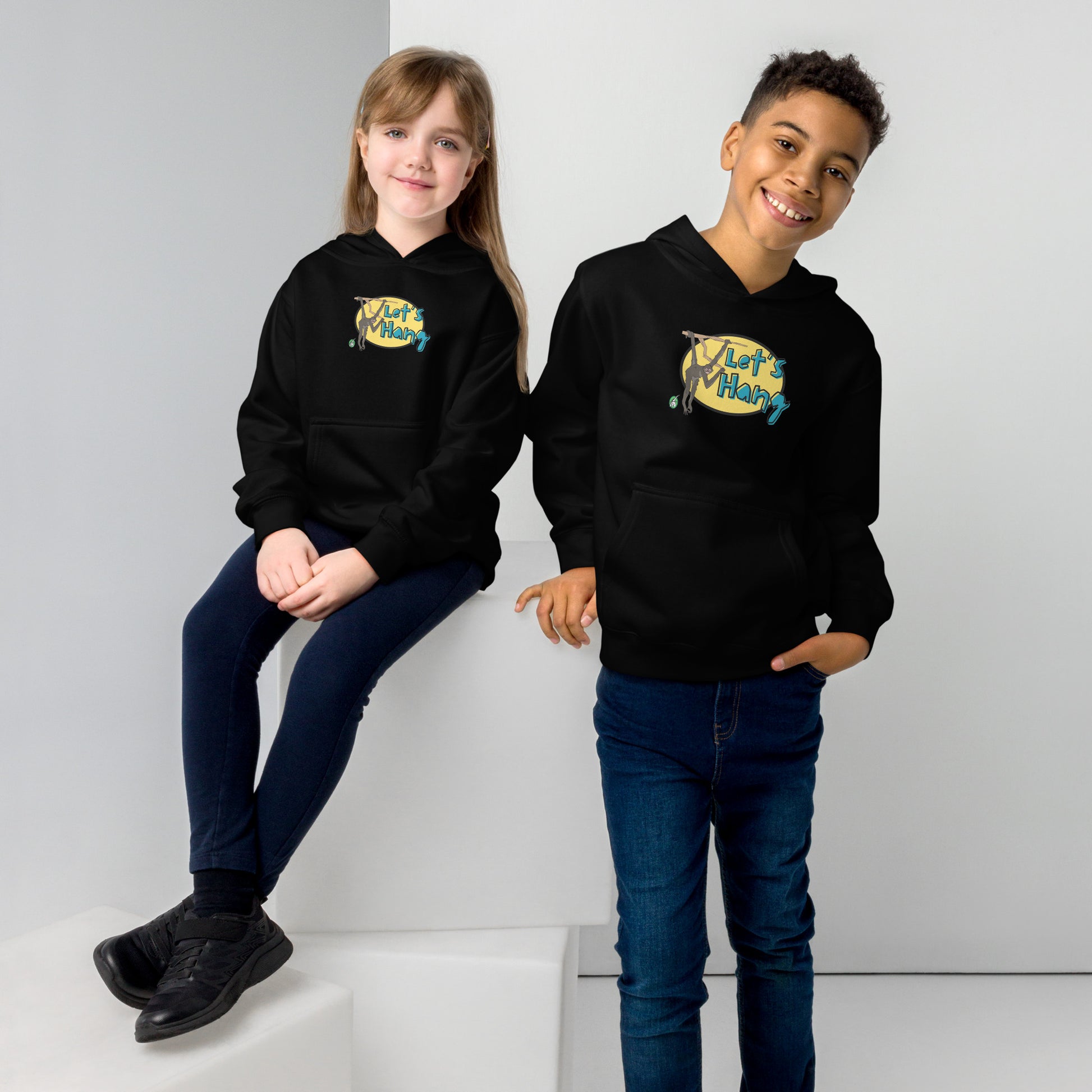 A boy standing and a girl sitting wearing a black hoodie with a funny graphic on the front of a monkey hanging from the tree branch saying, Let's Hang by Wizard Prints and jeans on a grey background