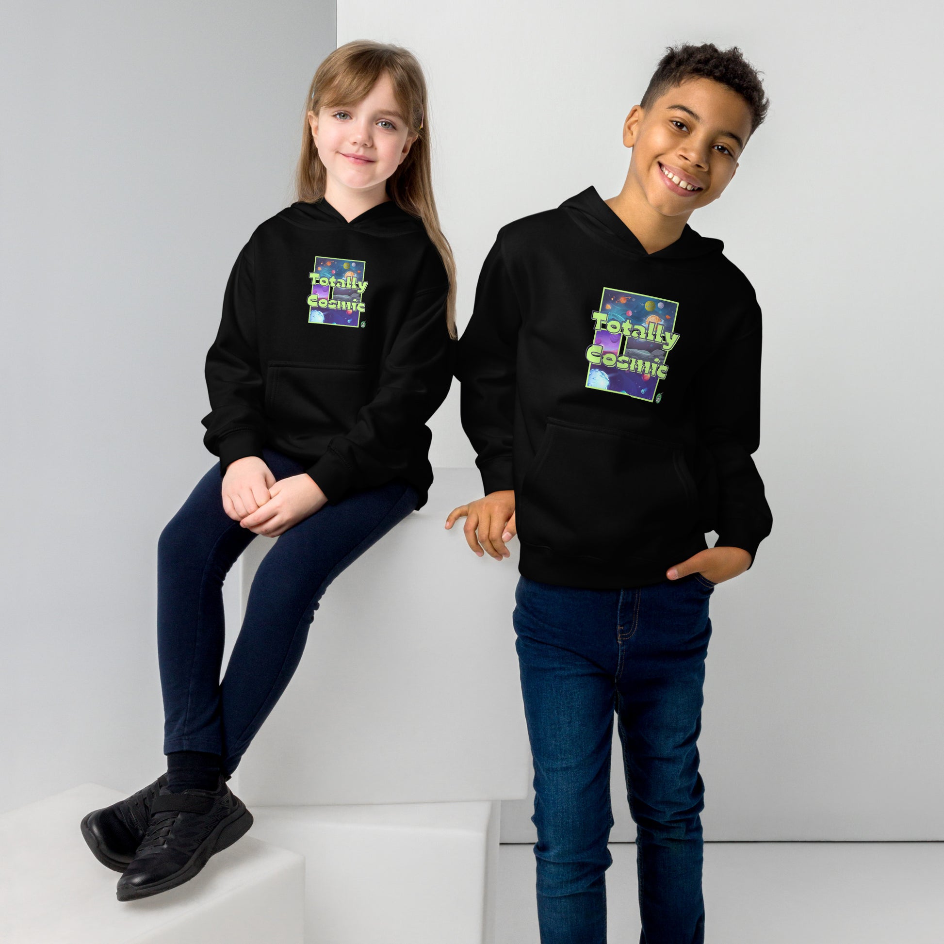A boy standing and a girl sitting both wearing a black kids hoodie with a graphic of the cosmos and the words Totally Cosmic, by Wizard Prints
