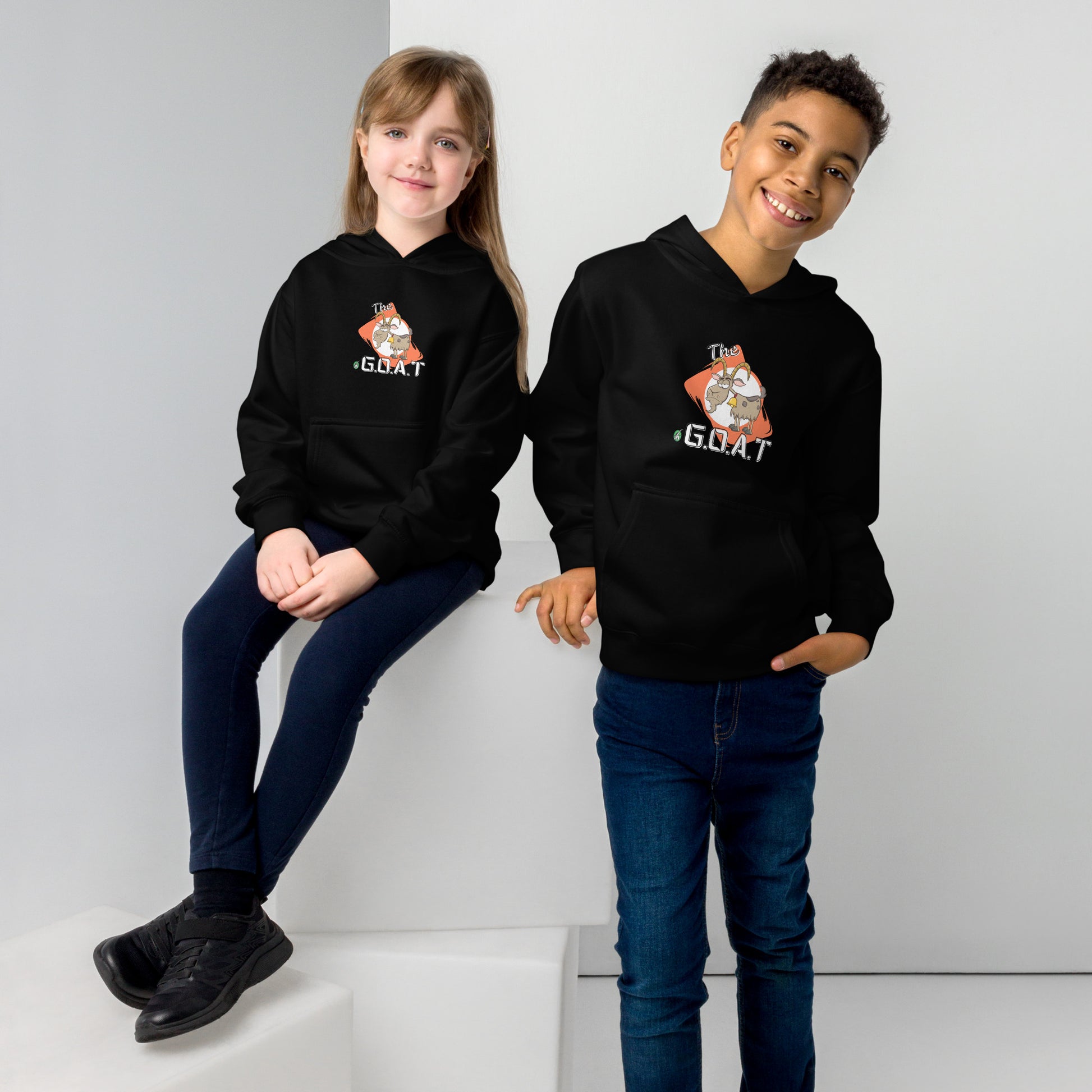 One boy standing and one girl sitting wearing a black kids hoodie with a graphic of a goat with the word G.O.A.T as in Greatest of all Time by Wizard Prints and jeans on a grey background
