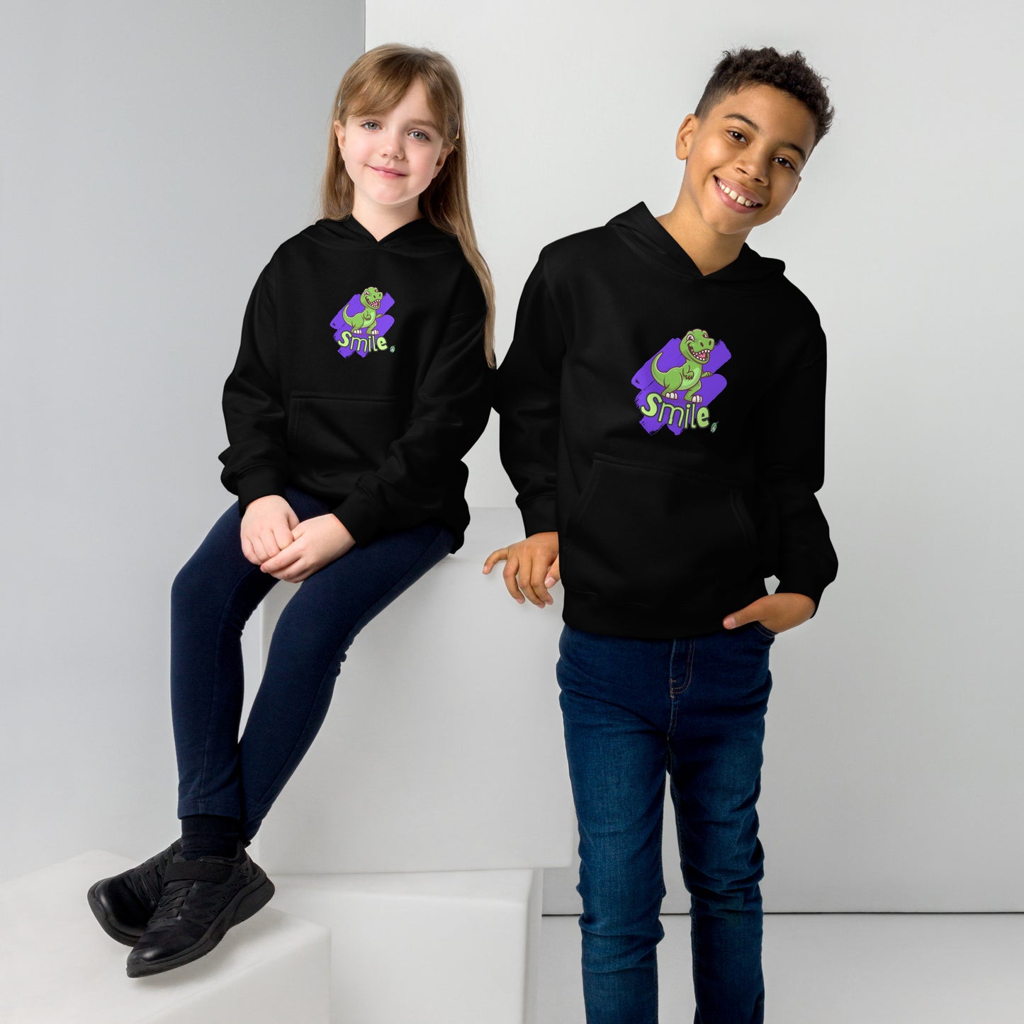 A boy standing and a girl sitting both wearing a black kids graphic hoodie with a green dinosaur and the word Smile on a purple background, by Wizard Prints
