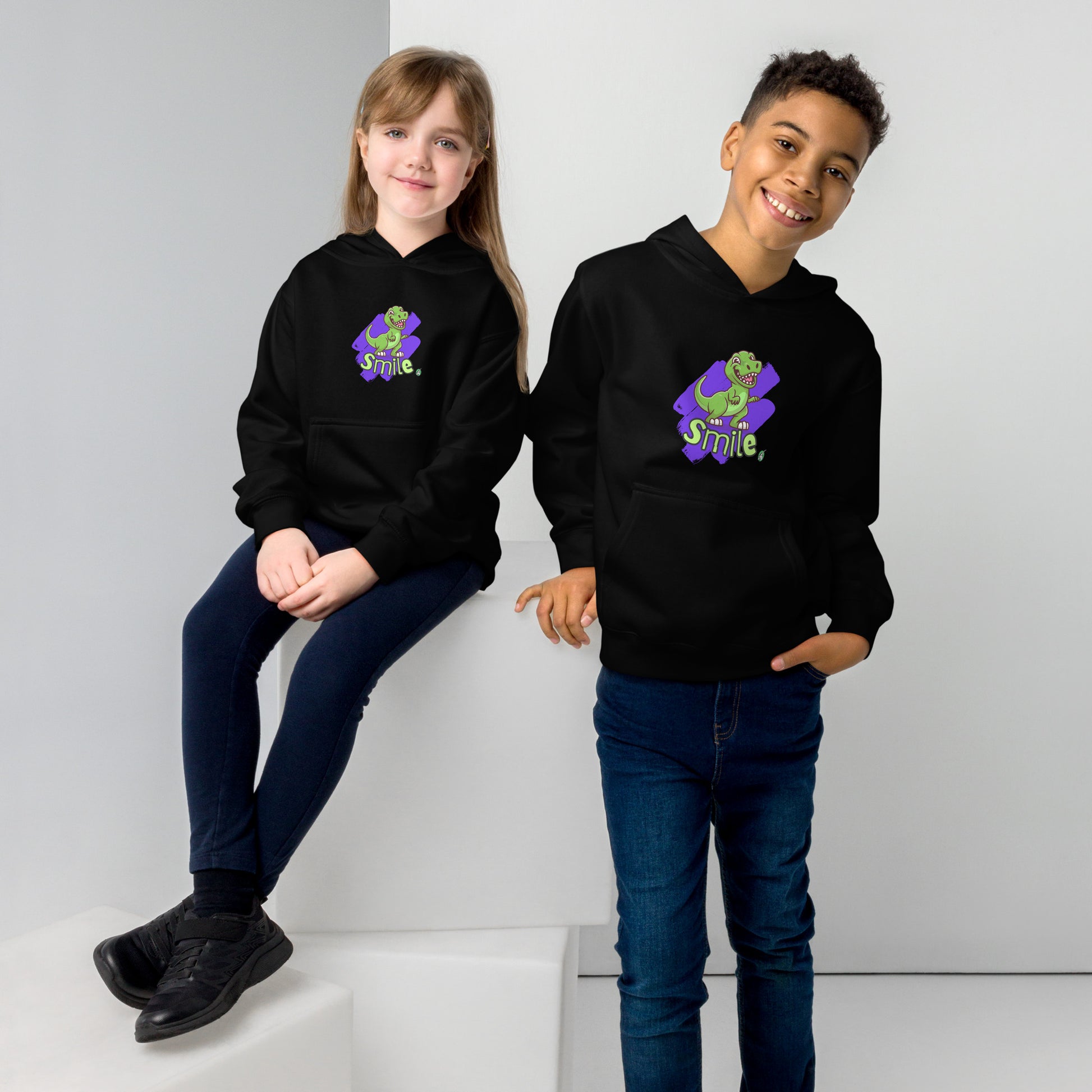 A boy standing and a girl sitting both wearing a black kids graphic hoodie with a green dinosaur and the word Smile on a purple background, by Wizard Prints