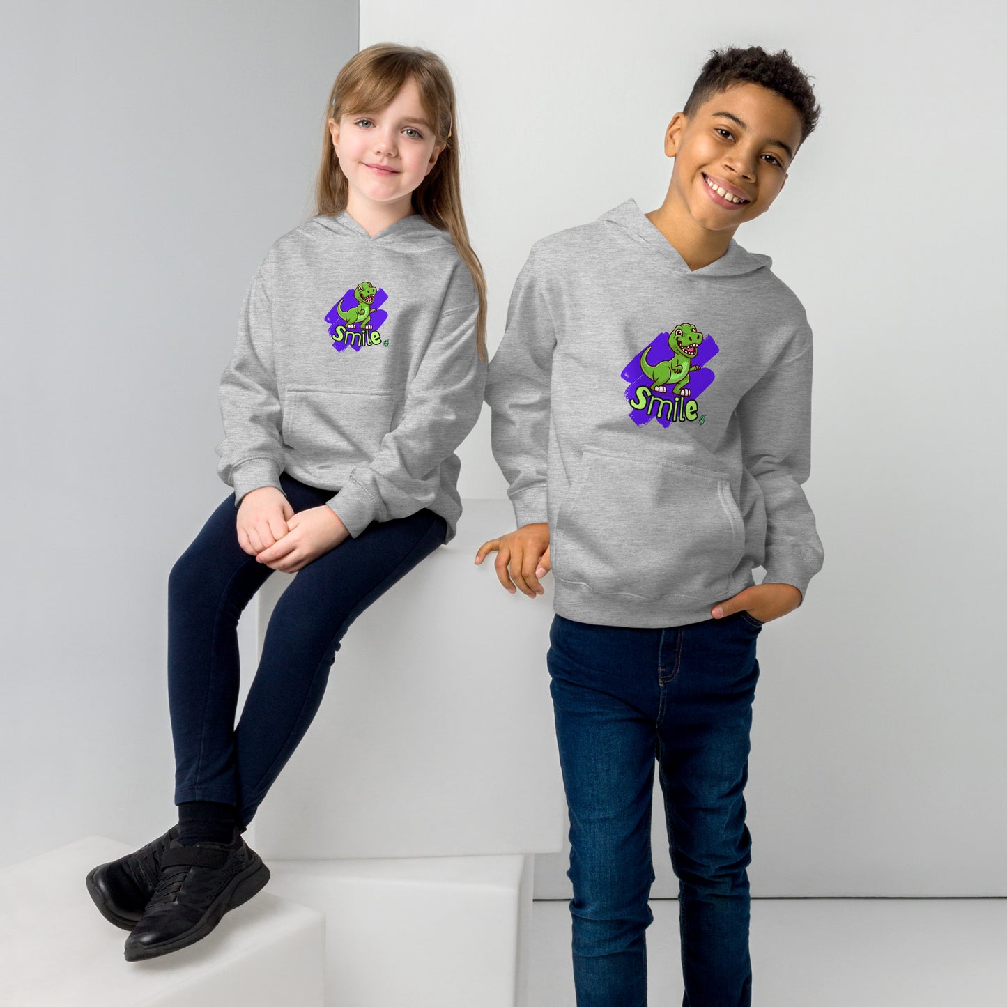 A boy standing and a girl sitting both wearing a grey kids graphic hoodie with a green dinosaur and the word Smile on a purple background, by Wizard Prints