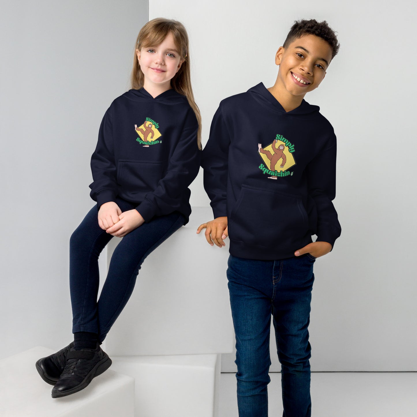 A boy standing and a girl sitting both wearing a navy blue kids graphic hoodie with a brown sasquatch and the statement, Simply Squatchin on a yellow background, by Wizard Prints