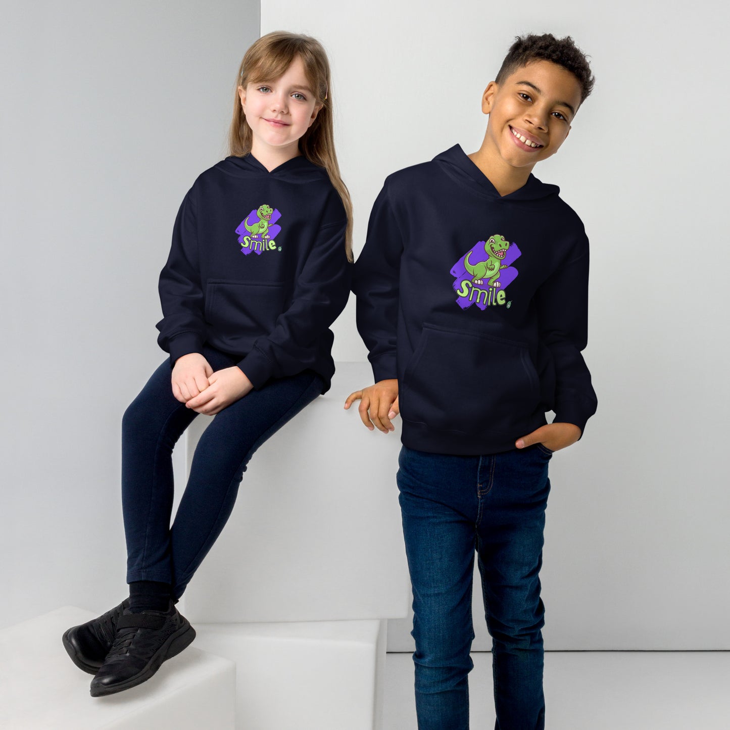 A boy standing and a girl sitting both wearing a navy blue kids graphic hoodie with a green dinosaur and the word Smile on a purple background, by Wizard Prints