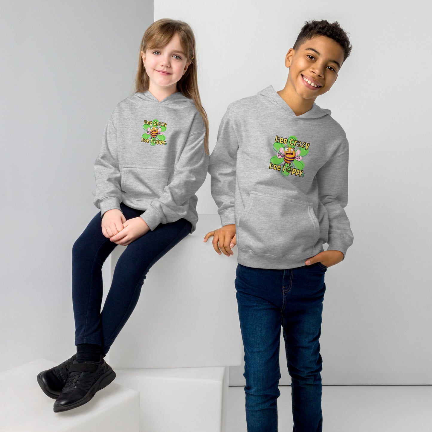  A boy standing and a girl sitting both wearing a grey kids hoodie with the printed graphic of a crazy bee and the statement, Bee Crazy Bee Happy. Designed by Wizard Prints