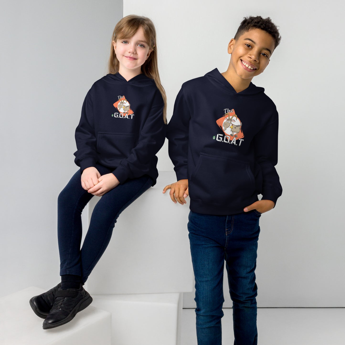 One boy standing and one girl sitting wearing a navy kids hoodie with a graphic of a goat with the word G.O.A.T as in Greatest of all Time by Wizard Prints and jeans on a grey background