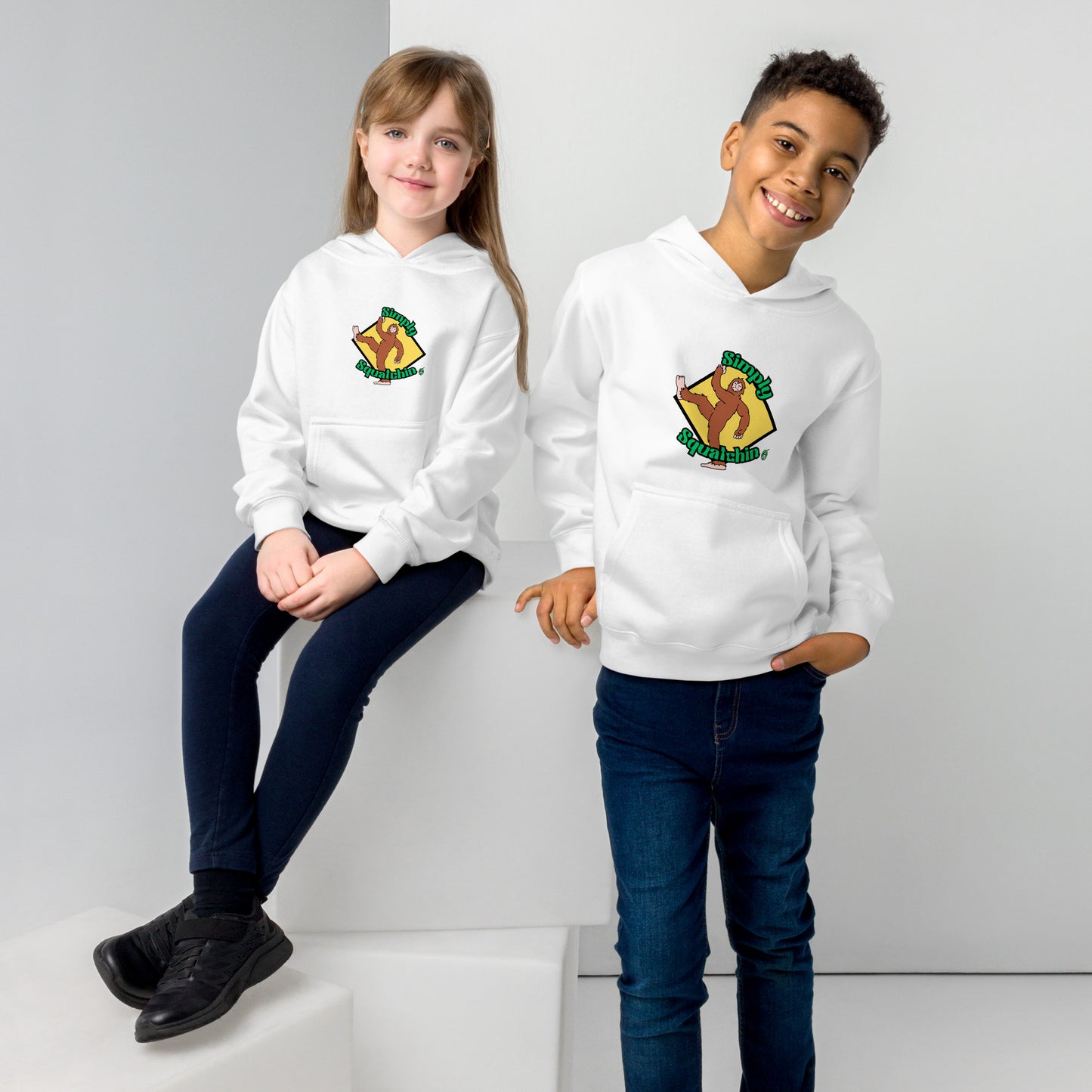 A girl sitting and a boy standing both wearing a white kids graphic hoodie with a brown sasquatch and the statement, Simply Squatchin on a yellow background, by Wizard Prints