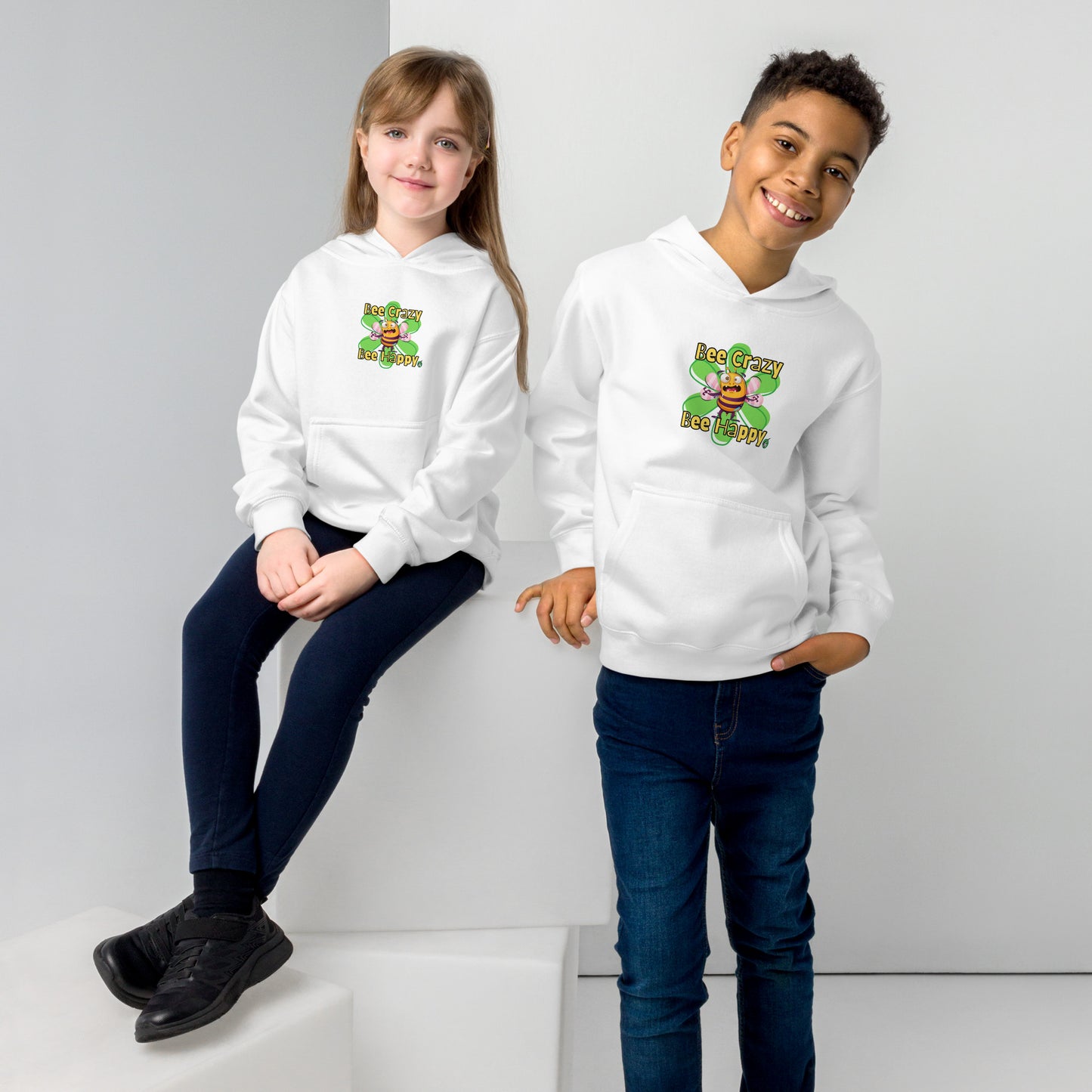  A boy standing and a girl sitting both wearing a white kids hoodie with the printed graphic of a crazy bee and the statement, Bee Crazy Bee Happy. Designed by Wizard Prints