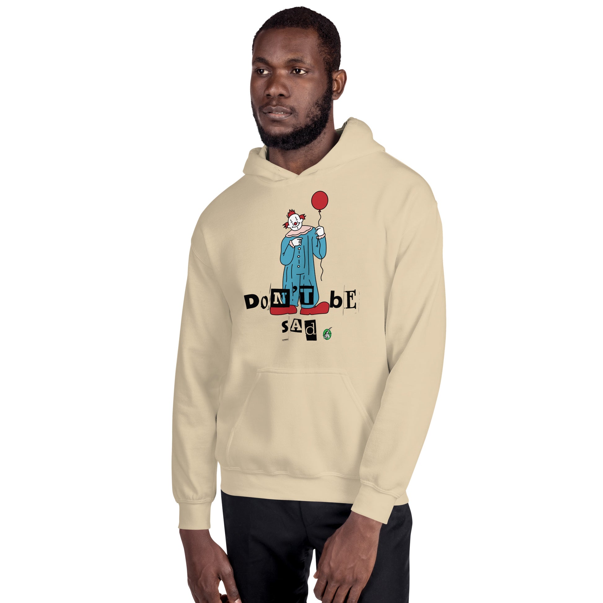 A man standing and wearing a sand beige hoodie with a printed graphic of a clown holding a balloon with the words, 'Don't Be Sad'. Designed by Wizard Prints.