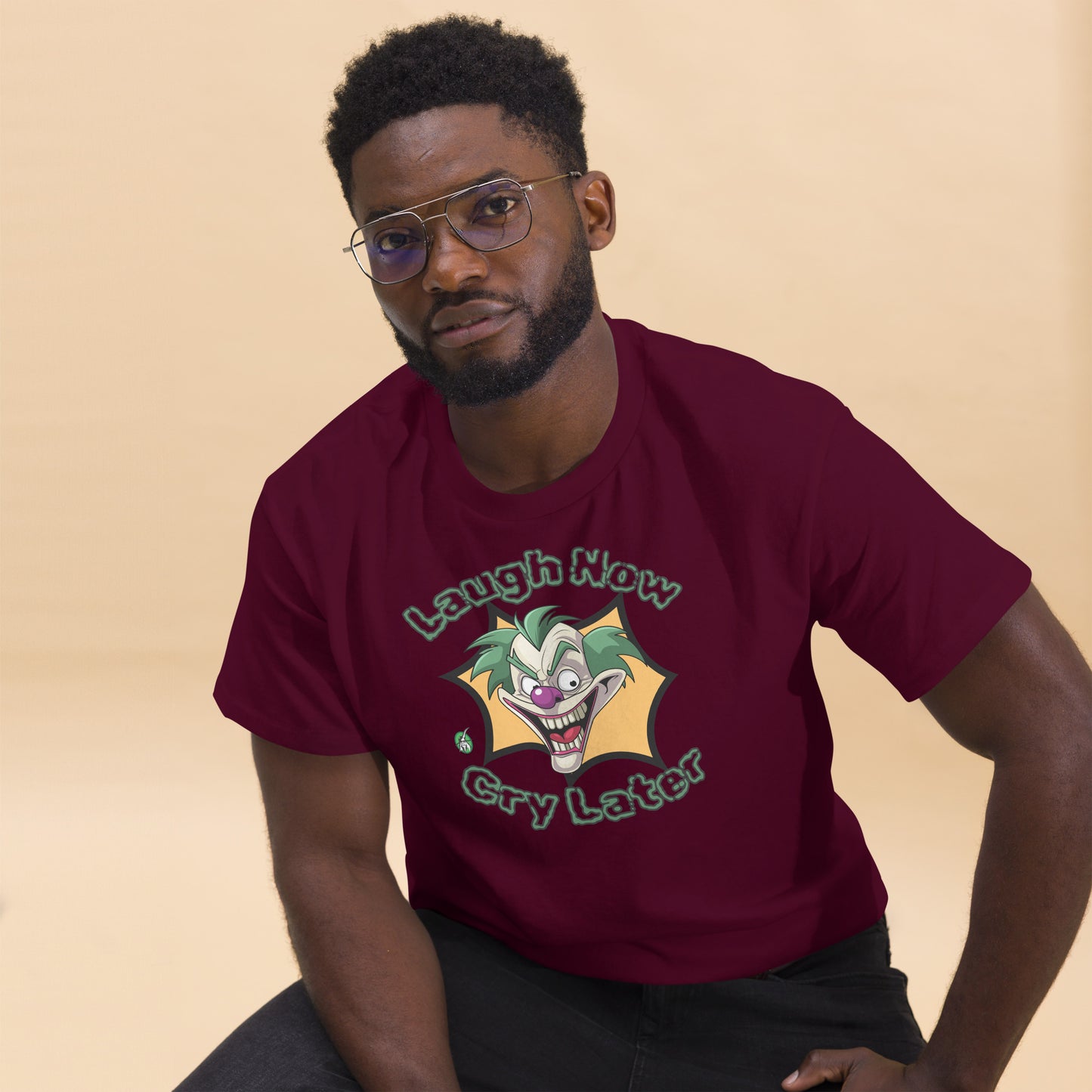 A man sitting wearing a maroon t-shirt with a printed graphic of a clown with the words, 'Laugh Now, Cry Later'. Designed by Wizard Prints.