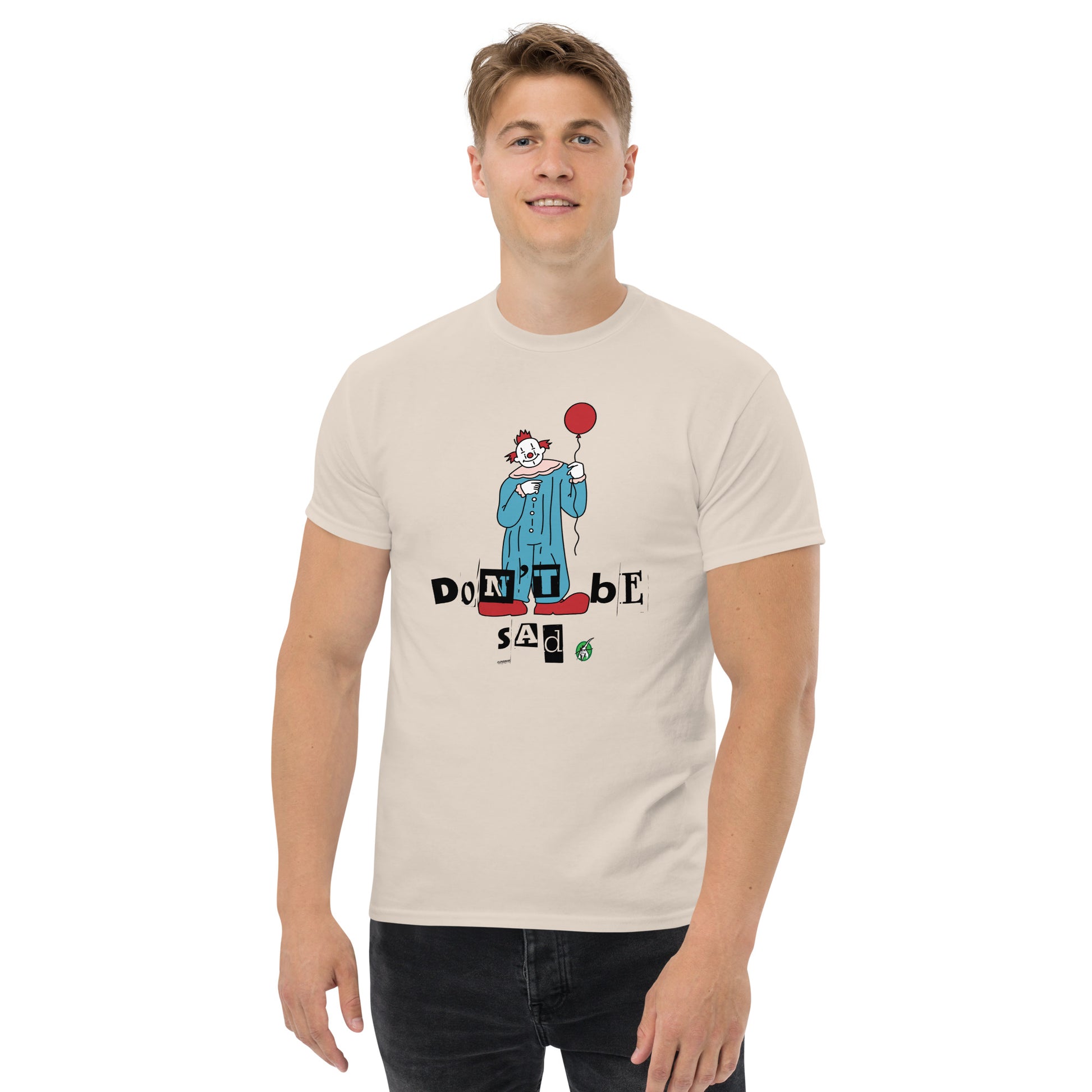 A man standing and wearing a beige t-shirt with a printed graphic of a clown holding a balloon with the words, 'Don't be sad'. Designed by Wizard Prints.