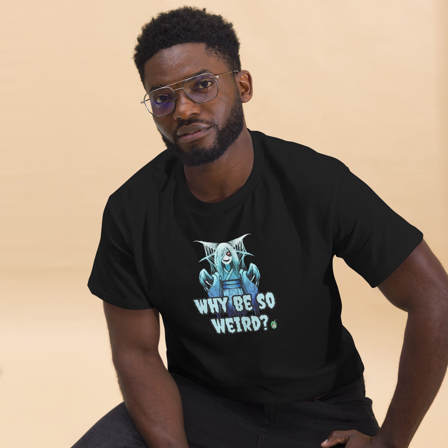 A man sitting and wearing a black t-shirt with a printed graphic of a blue ghost girl with the words, 'Why Be So Weird?'. Designed by Wizard Prints.
