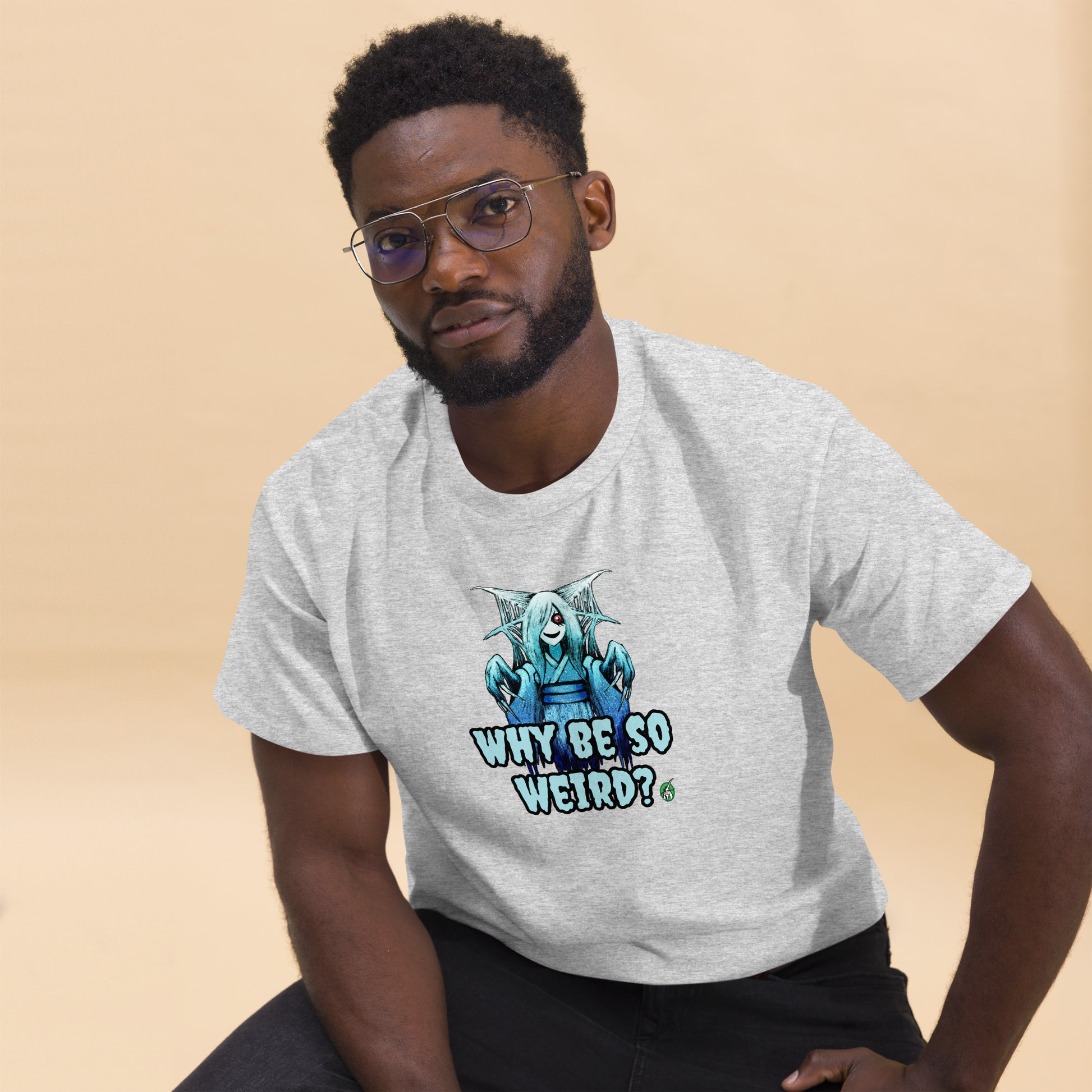 A man sitting and wearing a grey t-shirt with a printed graphic of a blue ghost girl with the words, 'Why Be So Weird?'. Designed by Wizard Prints.