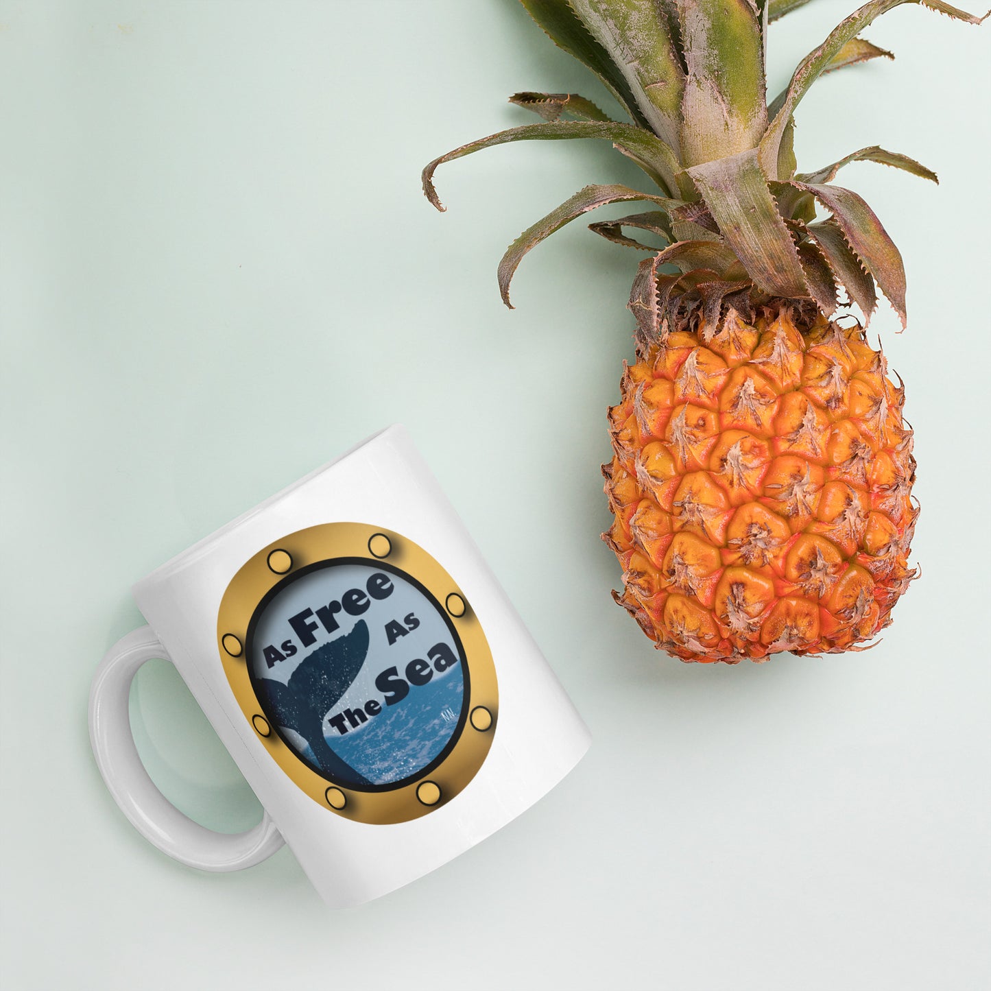 White mug with a whale and sea graphic and 'Free as the Sea' text  next to a pineapple on a light green background