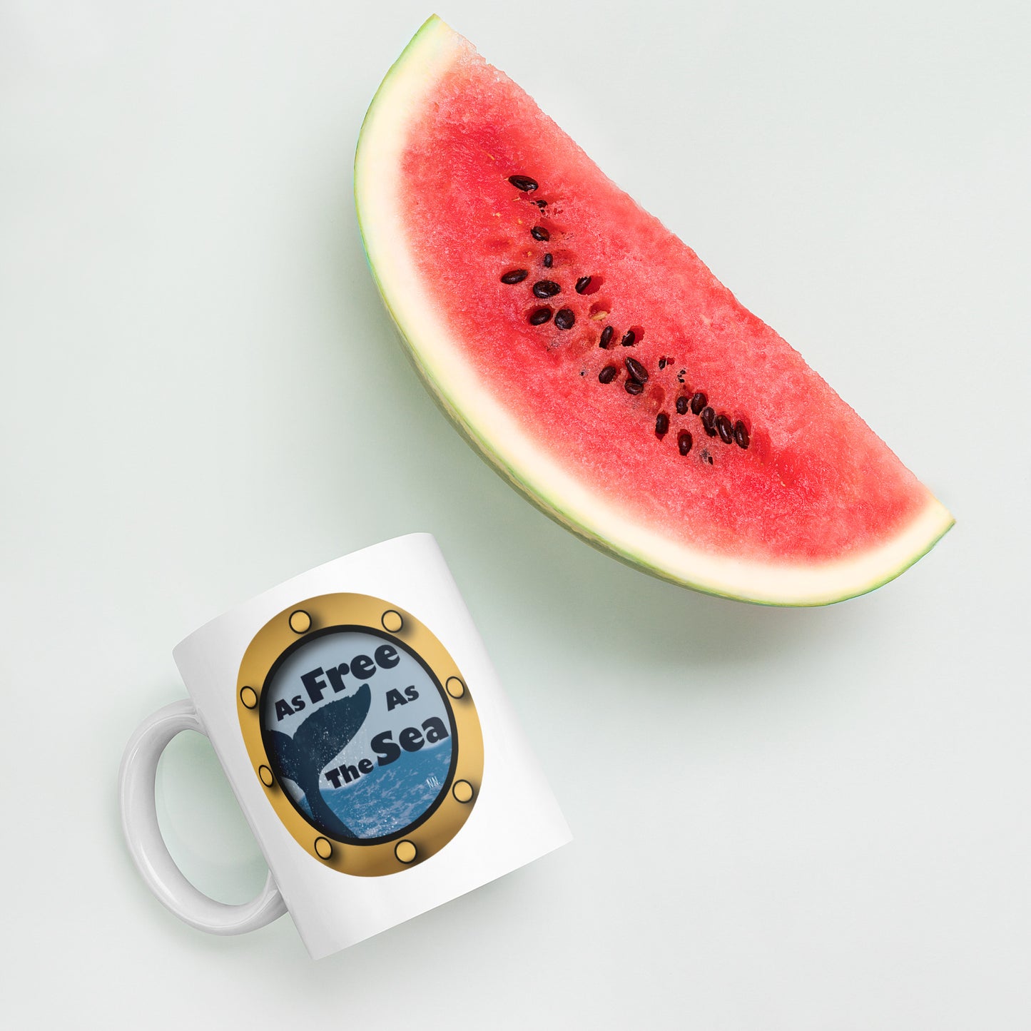 White mug with a whale and sea graphic and 'Free as the Sea' text  with decorative design next to a slice of watermelon on a light background