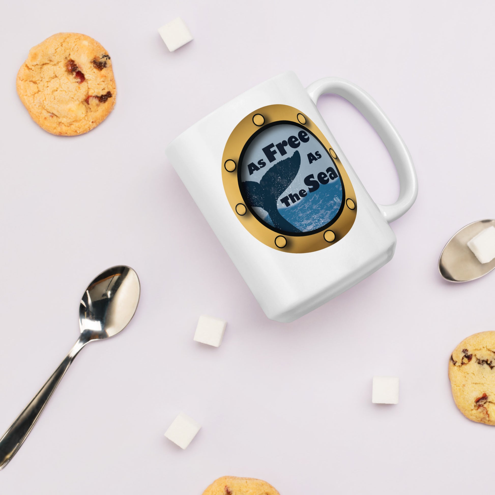 White mug with a whale and sea graphic and 'Free as the Sea' text on a light pink background with cookies and a spoon.