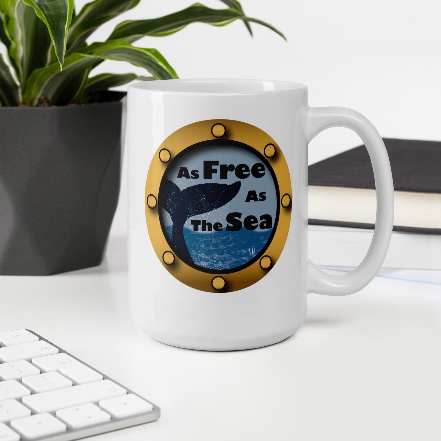 White mug with a whale and sea graphic and 'Free as the Sea' text on a desk with a plant and books.