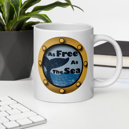 White mug with a whale and sea graphic and 'Free as the Sea' text on a desk with a plant and books in the background.