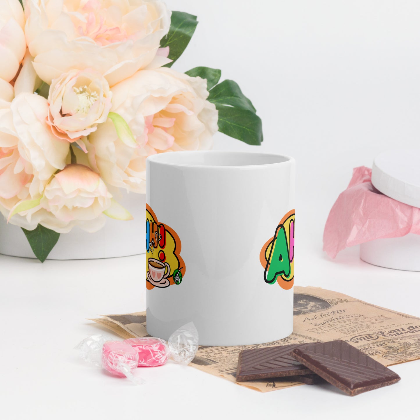 White mug with colourful design featuring a coffee cup and 'Ahhh!' text, placed on a surface with flowers and snacks.
