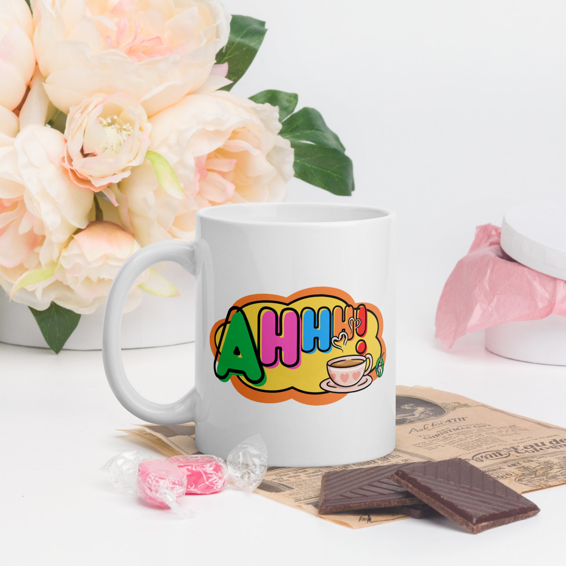 White mug with colourful design featuring a coffee cup and 'Ahhh!' text, placed on a surface with flowers and snacks.