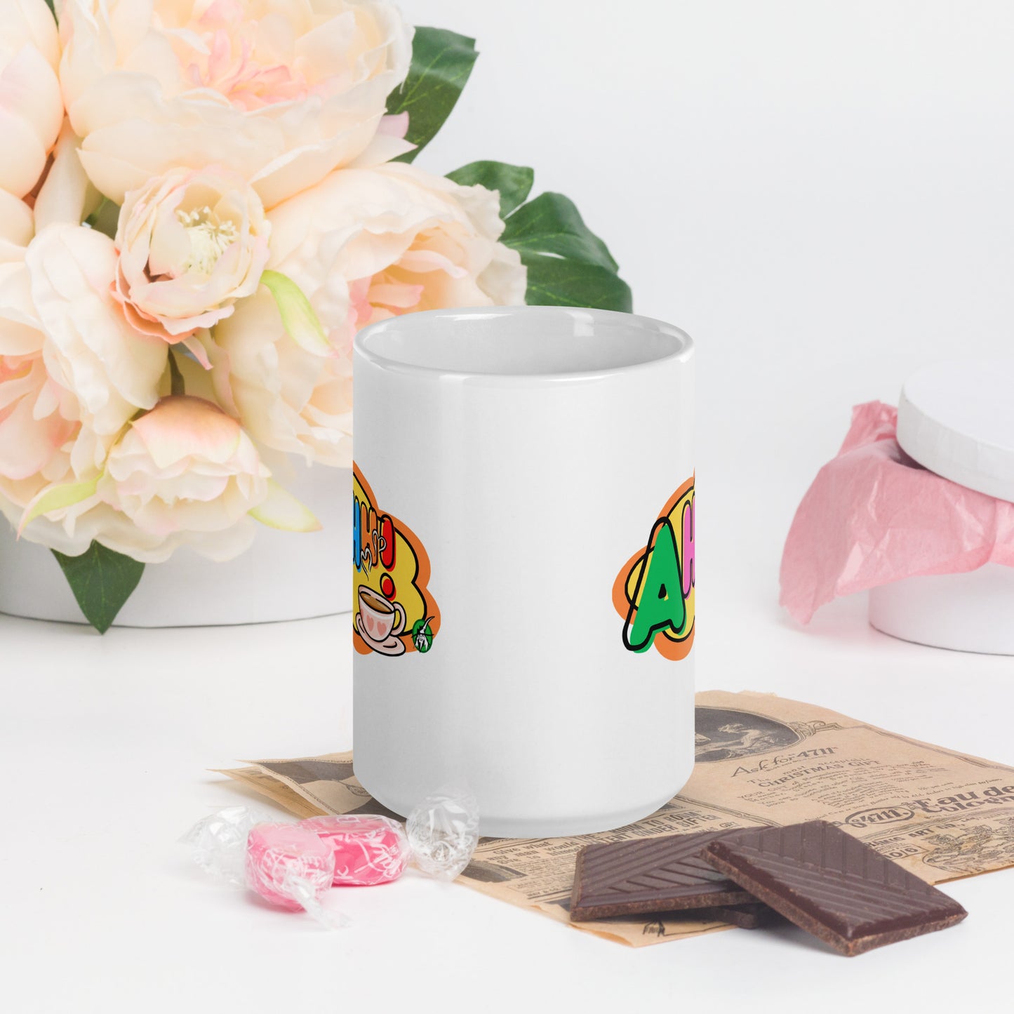 White mug with colourful design featuring a coffee cup and 'Ahhh!' text, placed on a surface with flowers and snacks.
