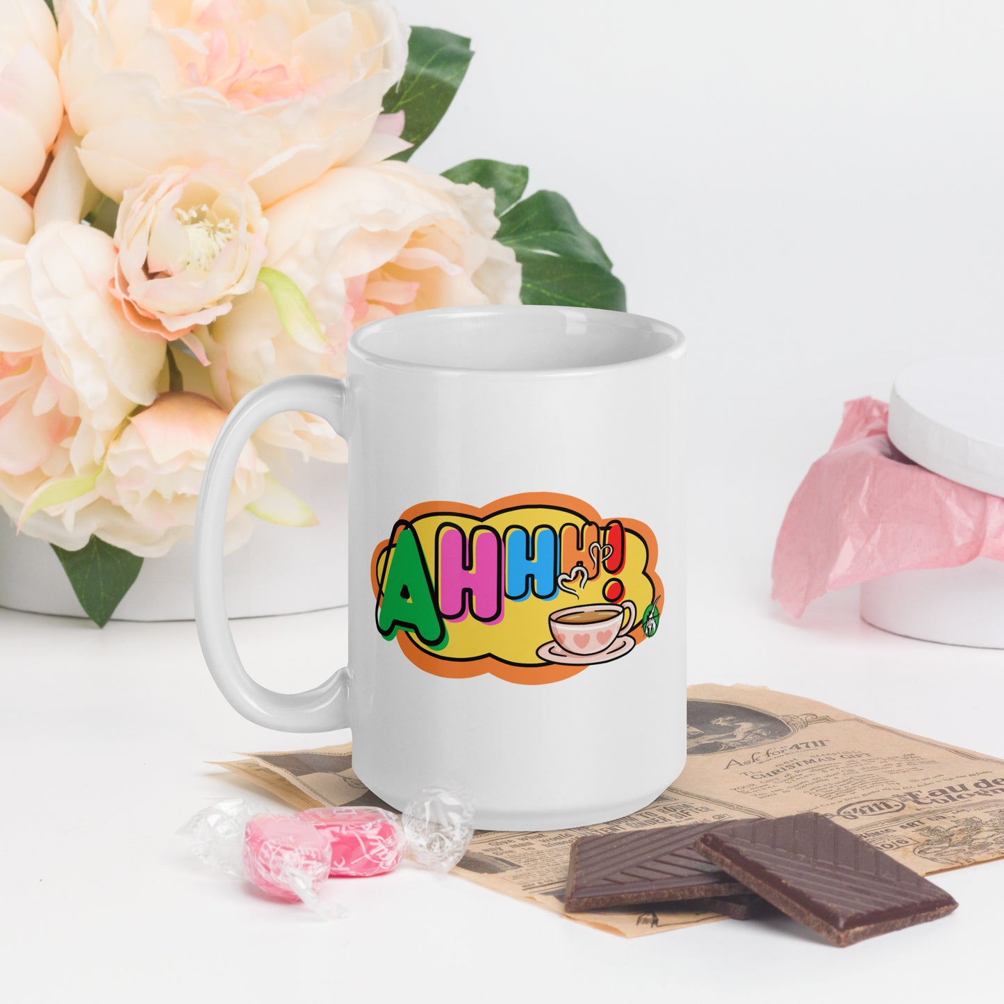 White mug with colourful design featuring a coffee cup and 'Ahhh!' text, placed on a surface with flowers and snacks.