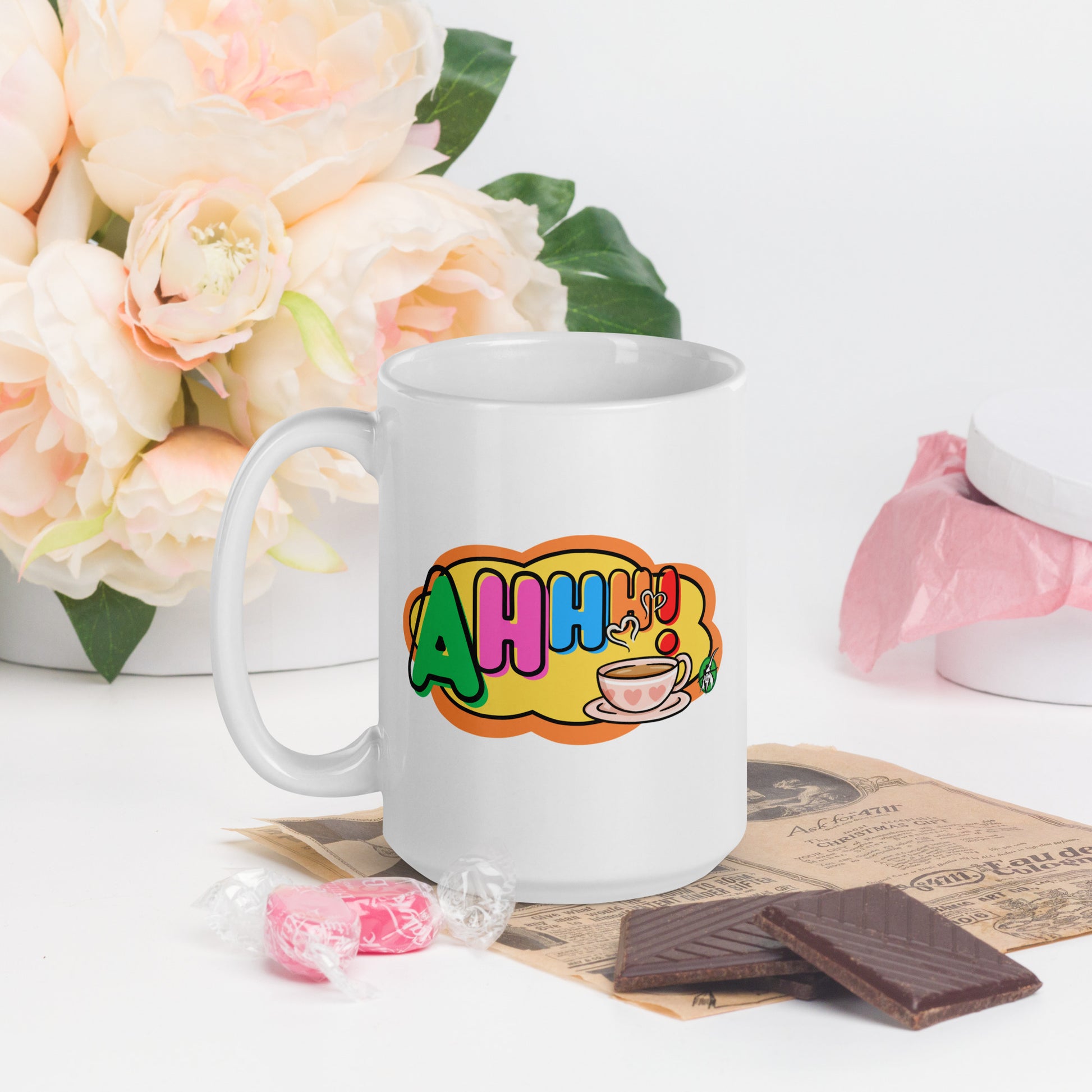 White mug with colourful design featuring a coffee cup and 'Ahhh!' text, placed on a surface with flowers and snacks.