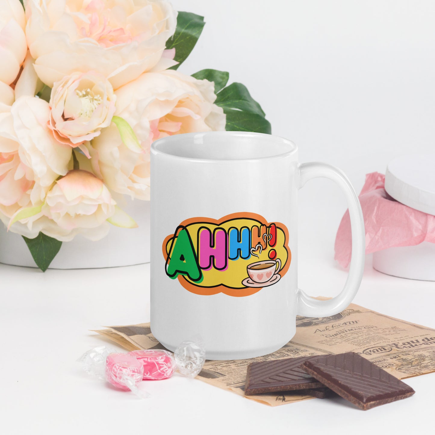 White mug with colourful design featuring a coffee cup and 'Ahhh!' text, placed on a surface with flowers and snacks.