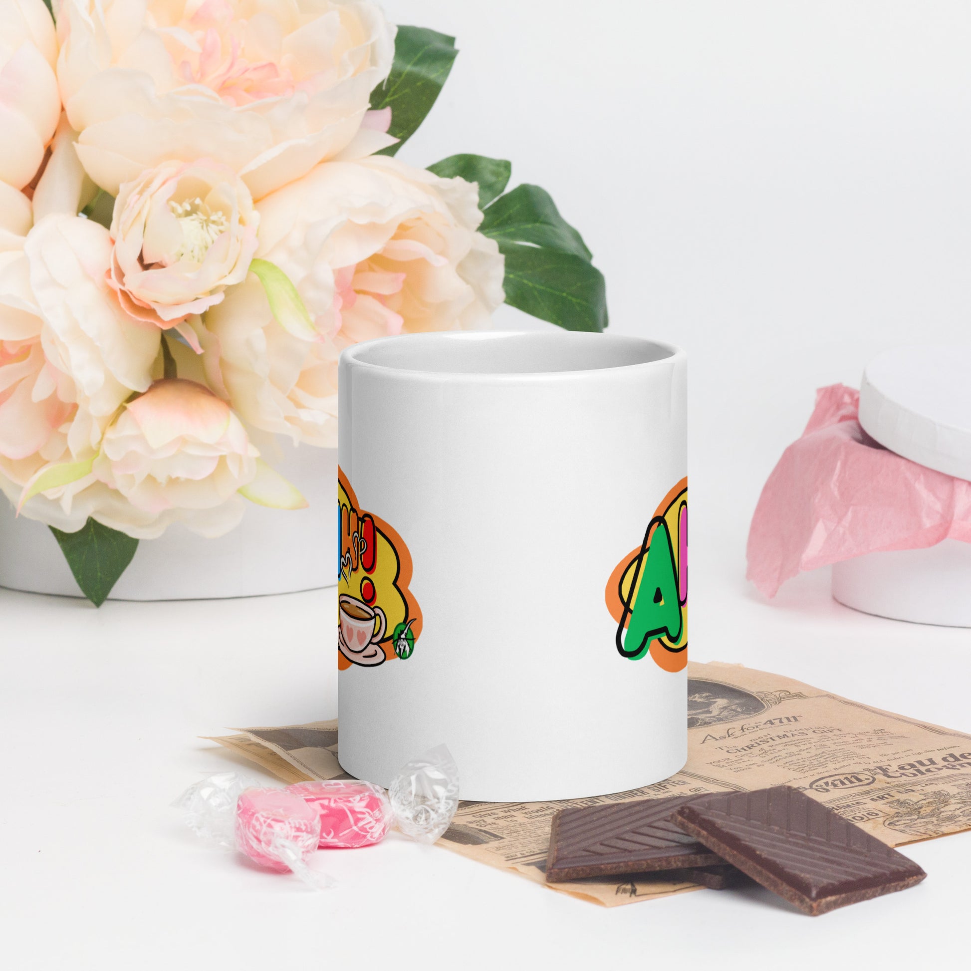 White mug with colourful design featuring a coffee cup and 'Ahhh!' text, placed on a surface with flowers and snacks.