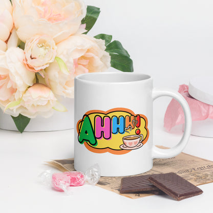 White mug with colourful design featuring a coffee cup and 'Ahhh!' text, placed on a surface with flowers and snacks.