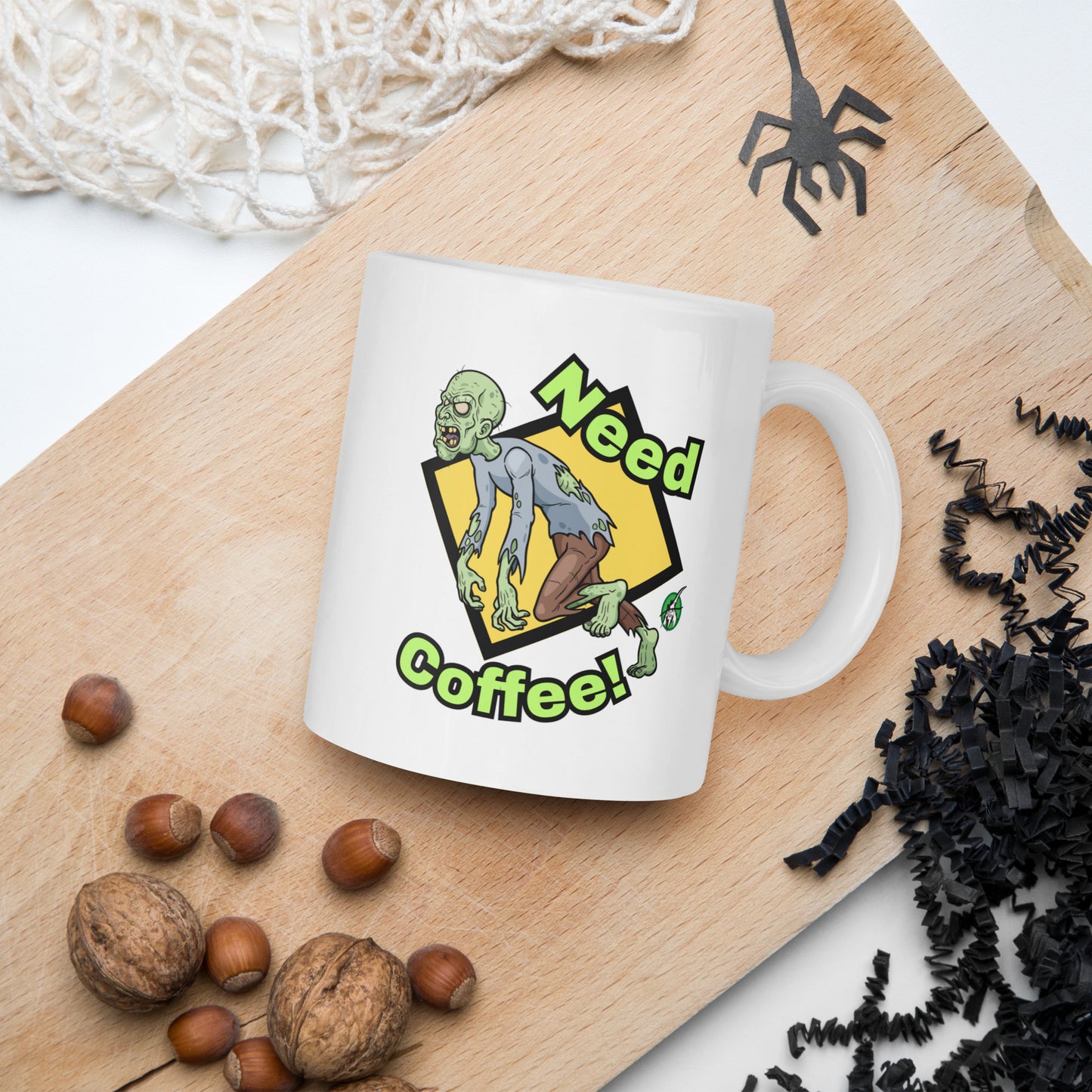 White mug with a zombie design and 'Need Coffee!' text on a wooden board with nuts and spider decorations.