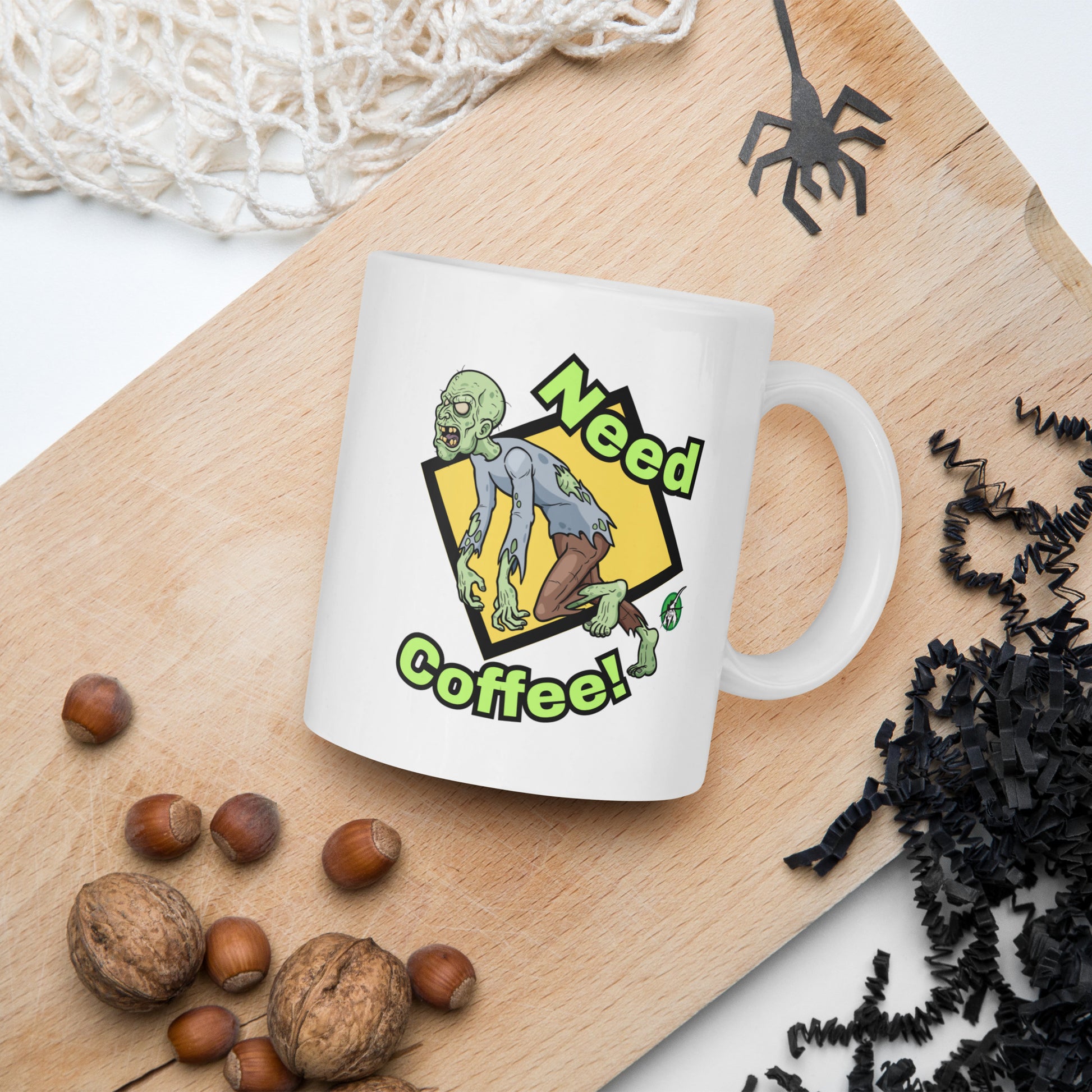 White mug with a zombie design and 'Need Coffee!' text on a wooden board with nuts and spider decorations.