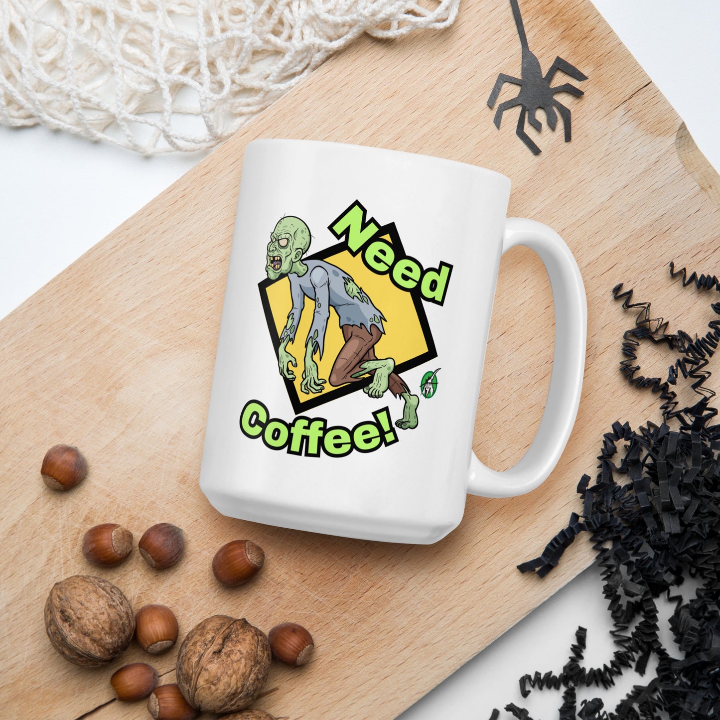 White mug with a zombie design and 'Need Coffee!' text on a wooden board with nuts and spider decorations.