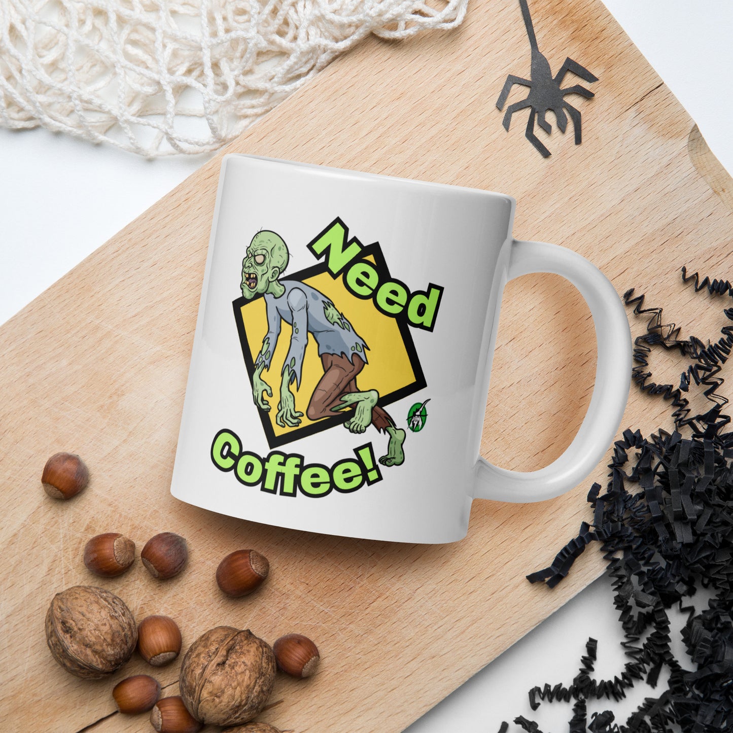 White mug with a zombie design and 'Need Coffee!' text on a wooden board with nuts and spider decorations.