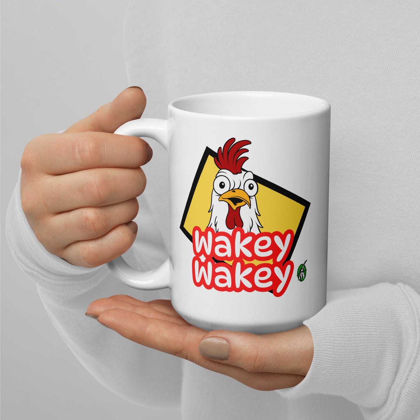 A Wizard Prints white mug with 'Wakey Wakey' and cartoon rooster graphic held by a person against a gray background.