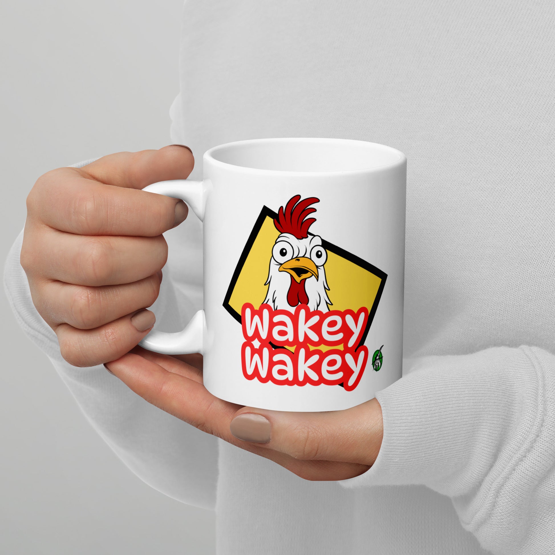 A Wizard Prints white mug with a cartoon rooster and 'Wakey Wakey' text held by a person against a light gray background.